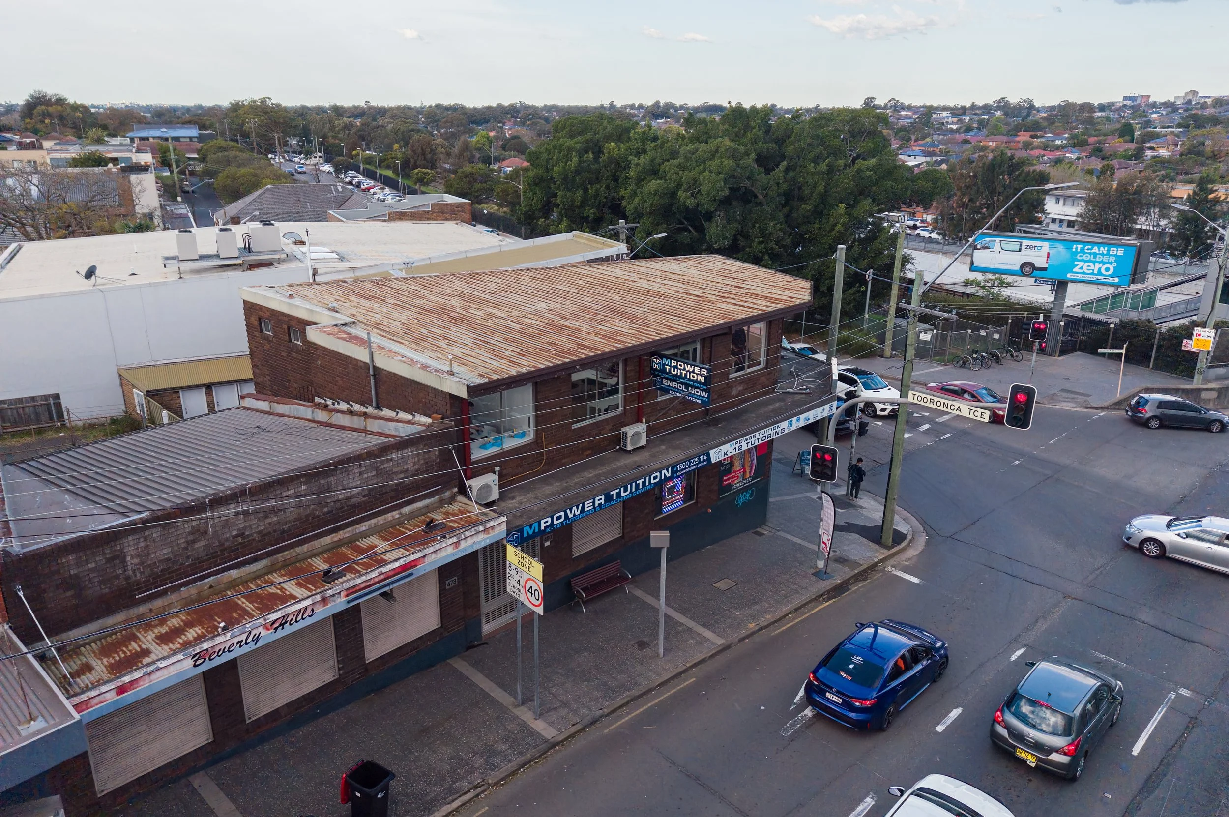 Street view showing an intersecting road with cars waiting at traffic lights, a brick building with signs for tutoring services, a billboard advertising a cold weather vehicle, and a background of trees and residential houses.