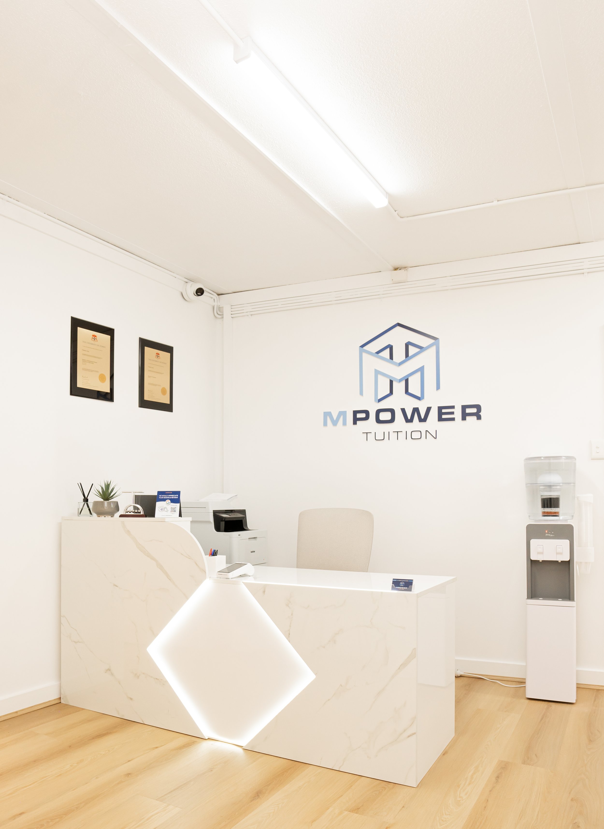 Modern reception area of M Power Tuition with a white marble desk, water dispenser, framed certificates on the wall, and the company logo on the wall.