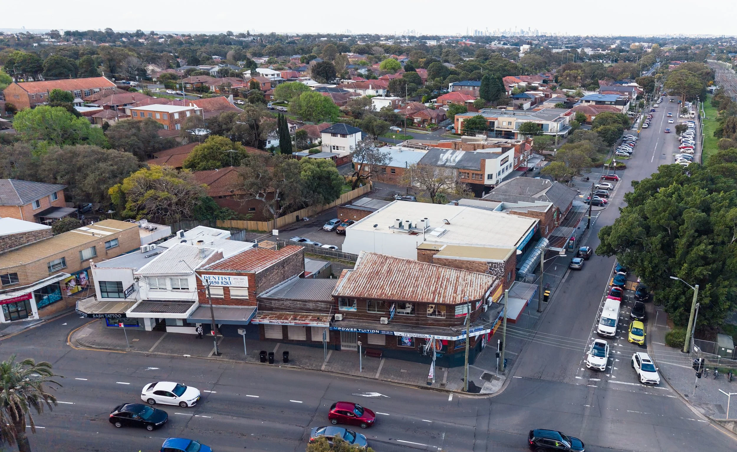 Aerial view of a suburban neighborhood with a main street lined with parked cars and small businesses, residential houses with trees and rooftops in the background, and city skyline in the distance.