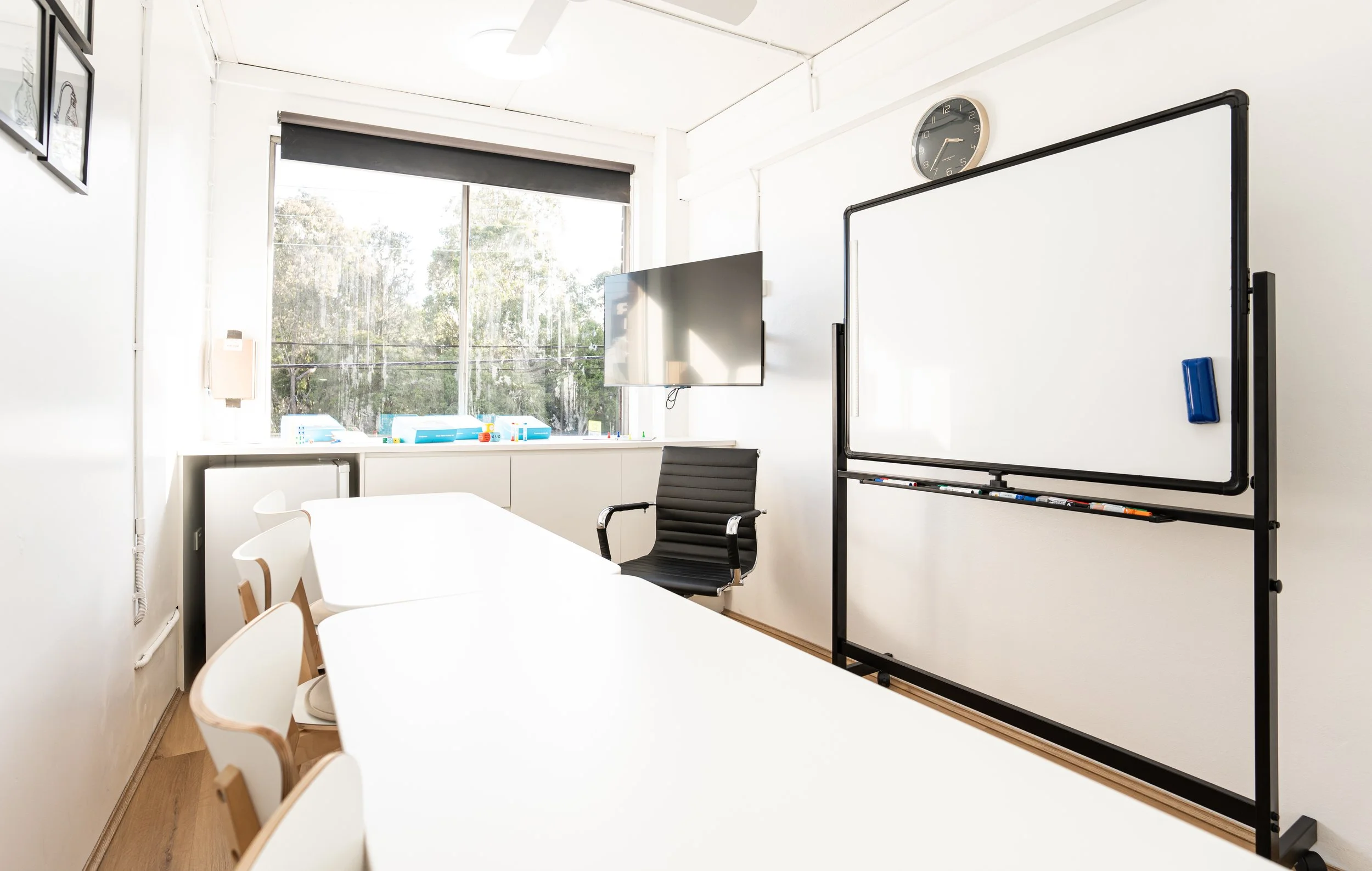 An empty conference room with a white table, white and wooden chairs, a wall-mounted TV, a whiteboard with markers, a wall clock, and a large window with trees outside.
