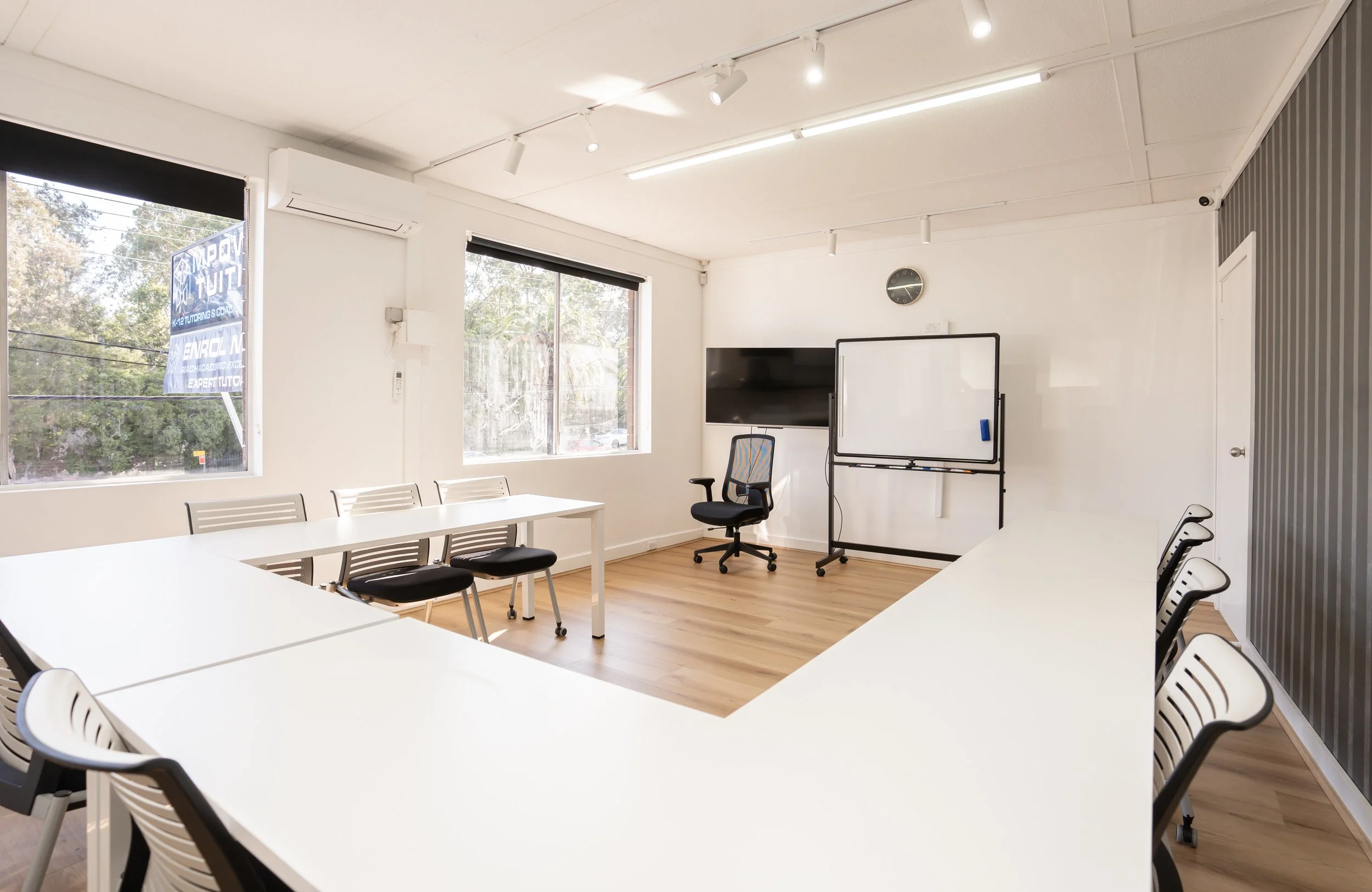 Empty conference room with white tables arranged in a U-shape, black chairs with white frames, a whiteboard on a stand, a wall-mounted TV, and a window showing trees outside.