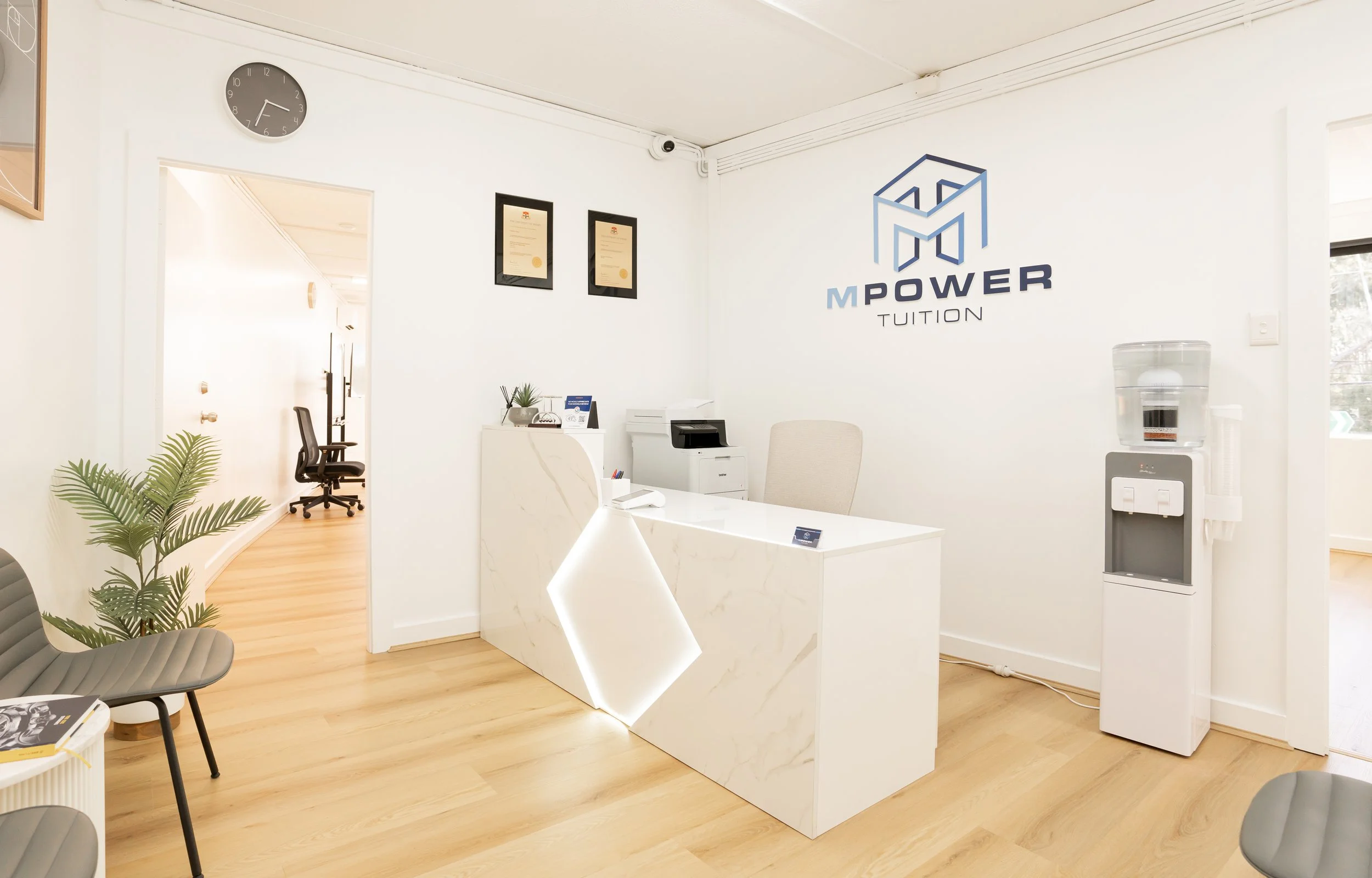 Modern office reception area with white walls, a white marble desk, a water cooler, framed certificates, a clock, and a wall logo for 'MPOWER TUITION.' there are office chairs, a potted plant, and a hallway visible through an open door.