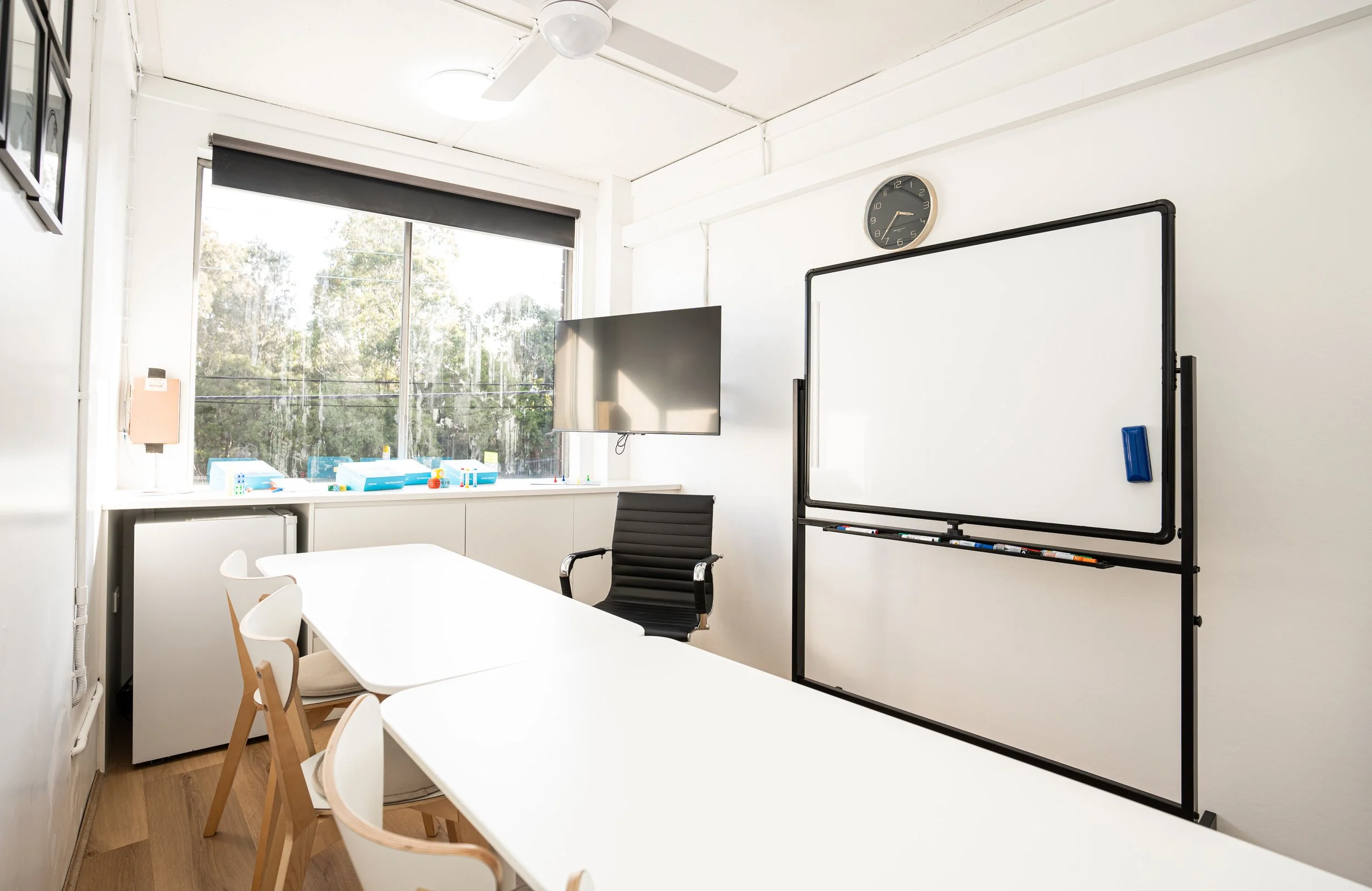 Empty conference room with white walls, a white table, white and wooden chairs, a whiteboard, a wall clock, and a television, with a large window showing trees outside.