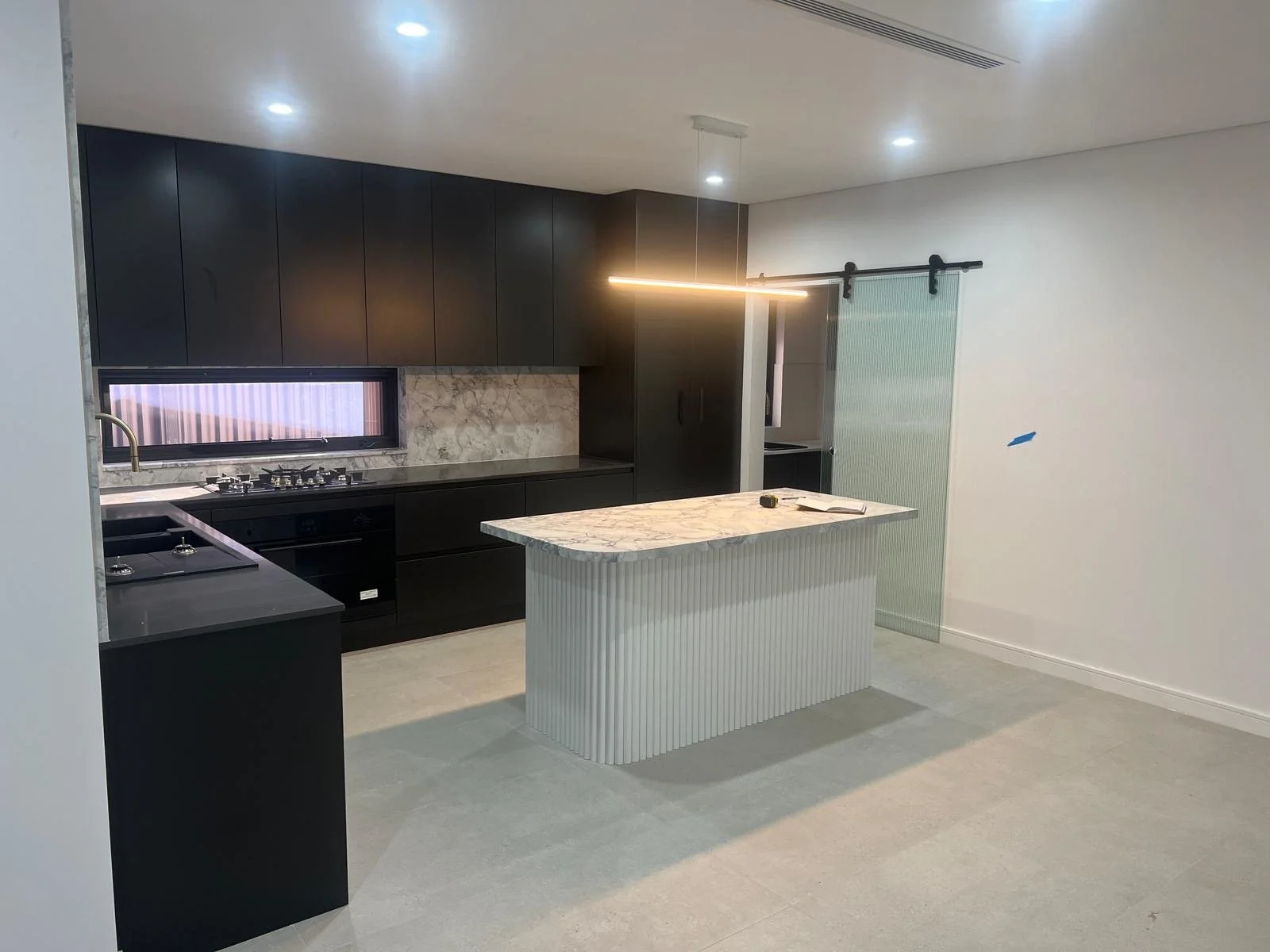 Modern kitchen with black cabinets, marble backsplash, marble island, black countertop, large window with striped blinds, ceiling lights, and a sliding door with frosted glass.