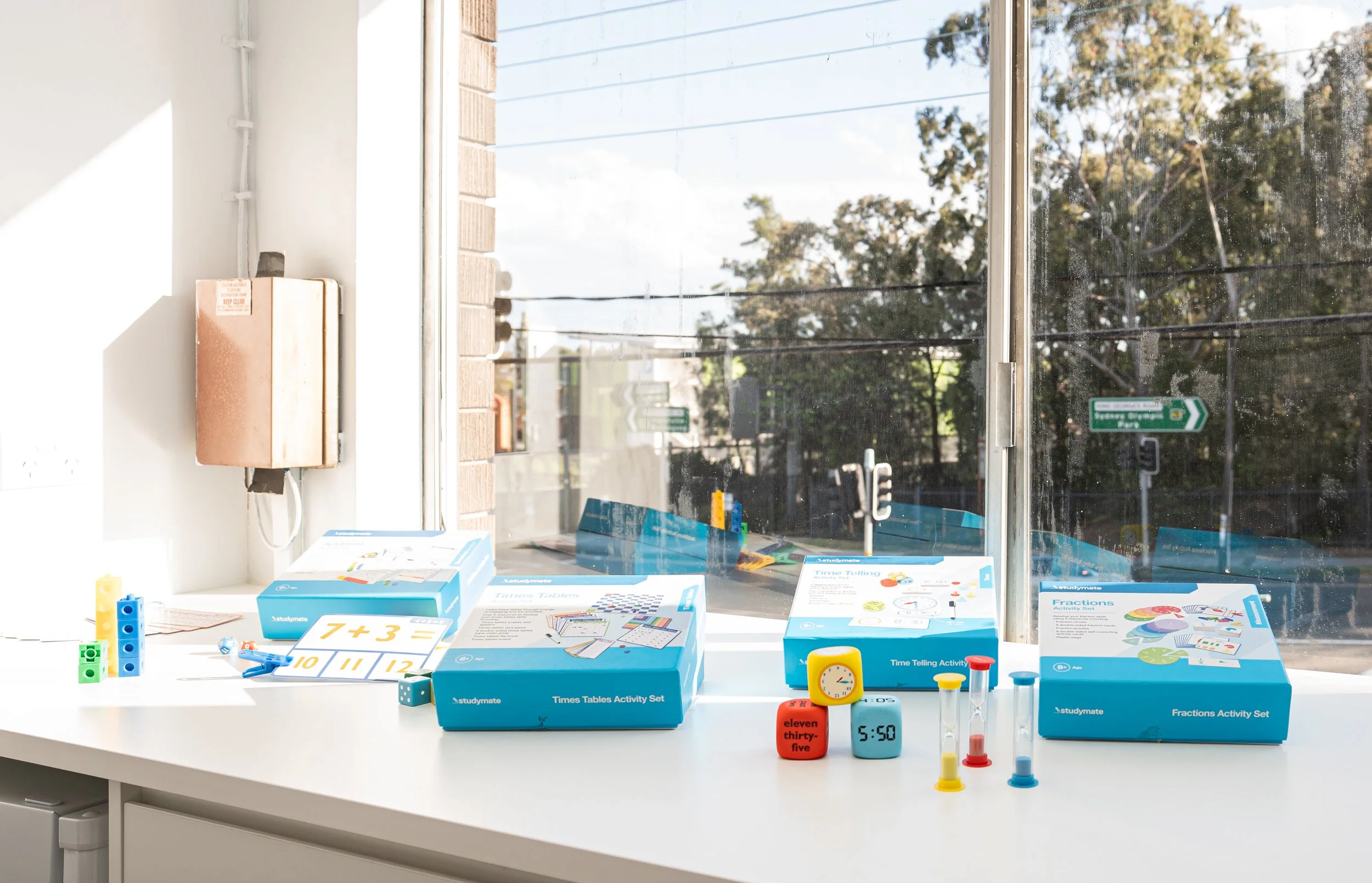 Educational math activity sets and tools on a white windowsill, including boxes labeled 'Times Tables Activity Set,' 'Time Telling Activity Set,' and 'Fractions Activity Set,' along with a digital clock, small hourglasses, colorful counting cubes, and educational flashcards.