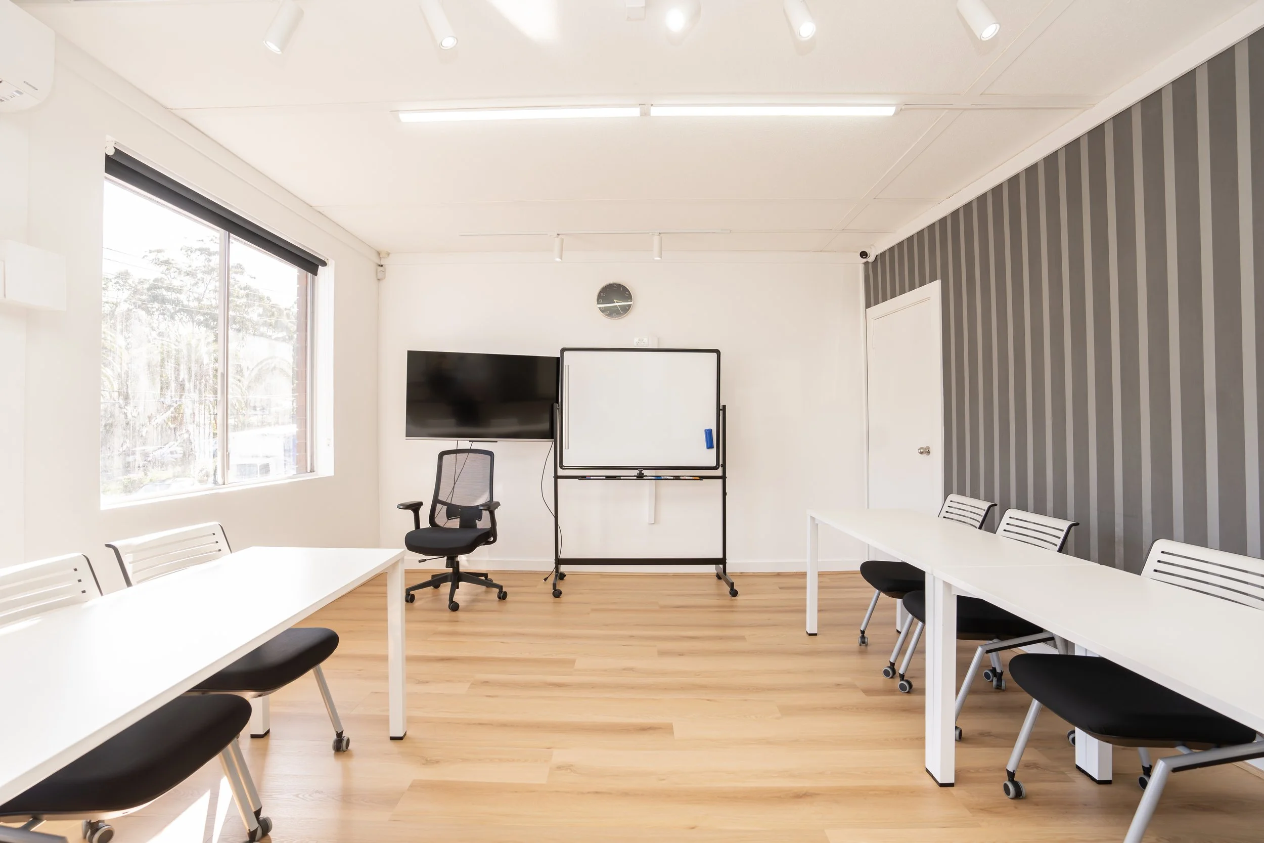 Empty classroom with white tables, black chairs, a whiteboard, a monitor, and a wall clock.