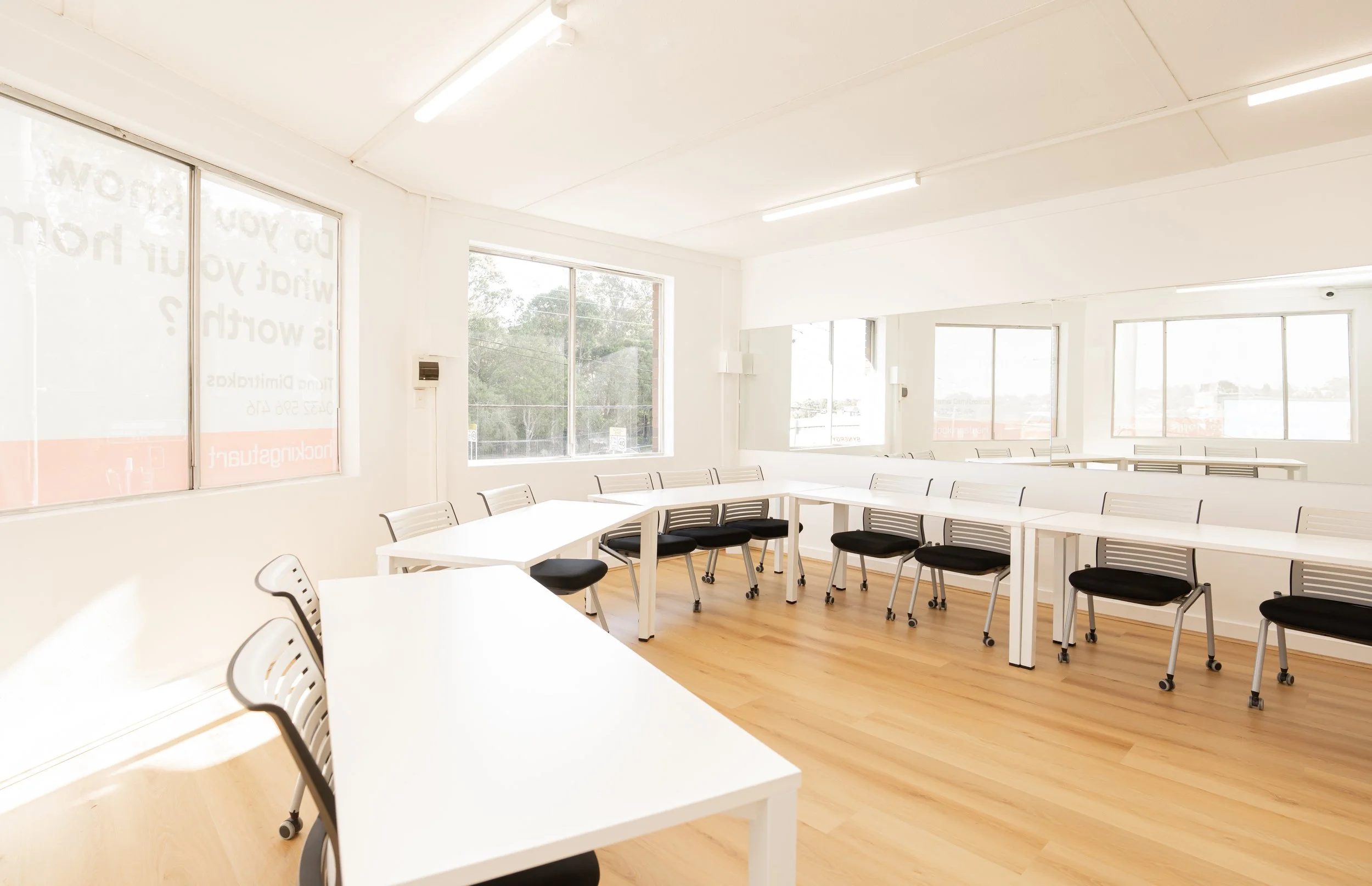 Empty classroom with white tables and black chairs, large windows, and wooden floor.