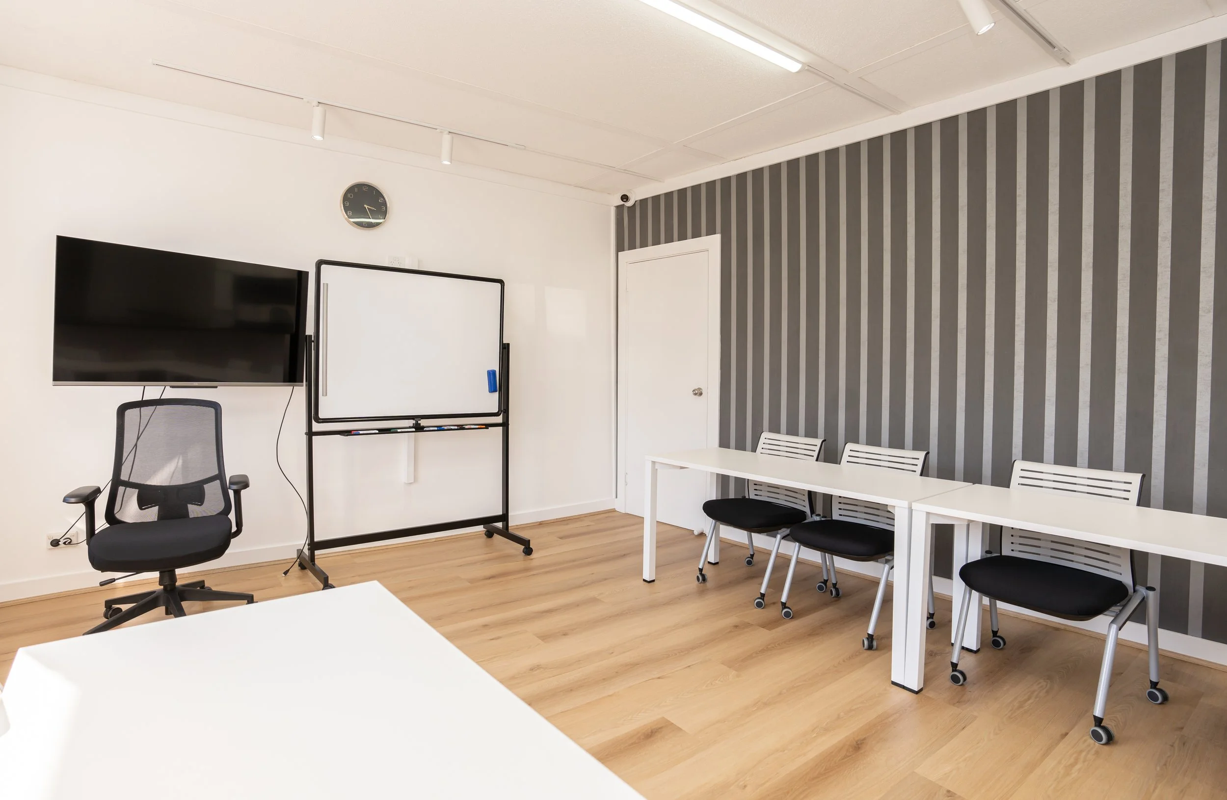 Empty classroom with white tables, black chairs, a whiteboard, a large monitor, a wall clock, and a gray striped accent wall.