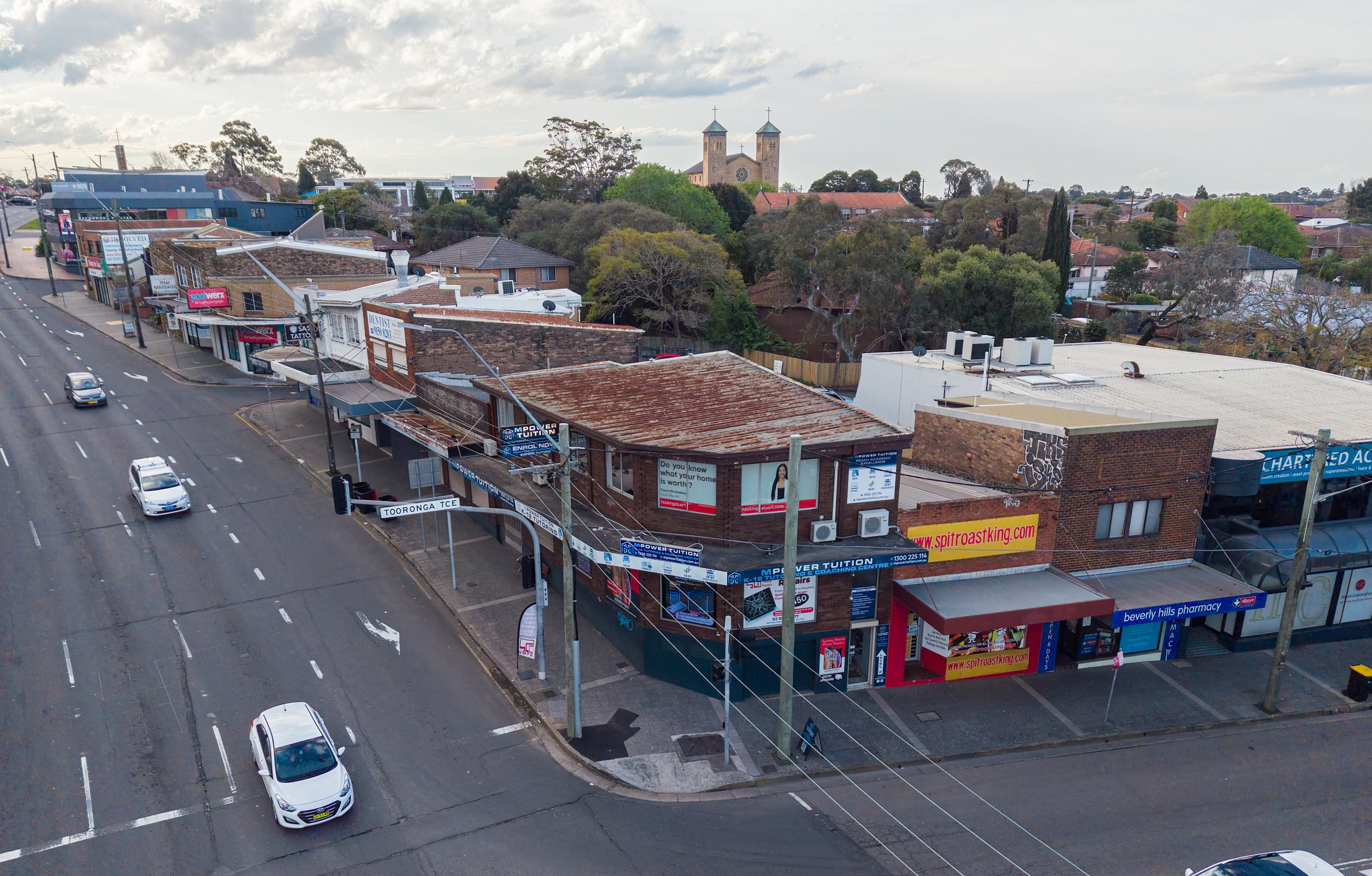 An aerial view of a small commercial area with multiple storefronts on a street, including a pharmacy and tutoring centers, with trees and residential buildings in the background.