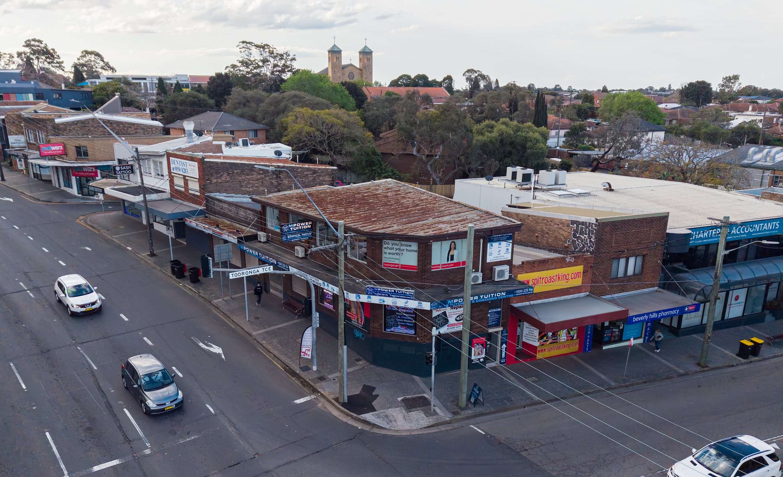 An aerial view of a small commercial street with shops, parked vehicles, utility poles, and some pedestrians. There are signs on the businesses, trees and a church with twin towers in the background.