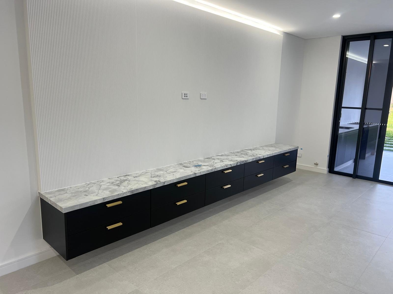A minimalist living room with a long black floating cabinet topped with white marble, set against a textured white wall, with a sliding glass door to the right leading to an outdoor space.