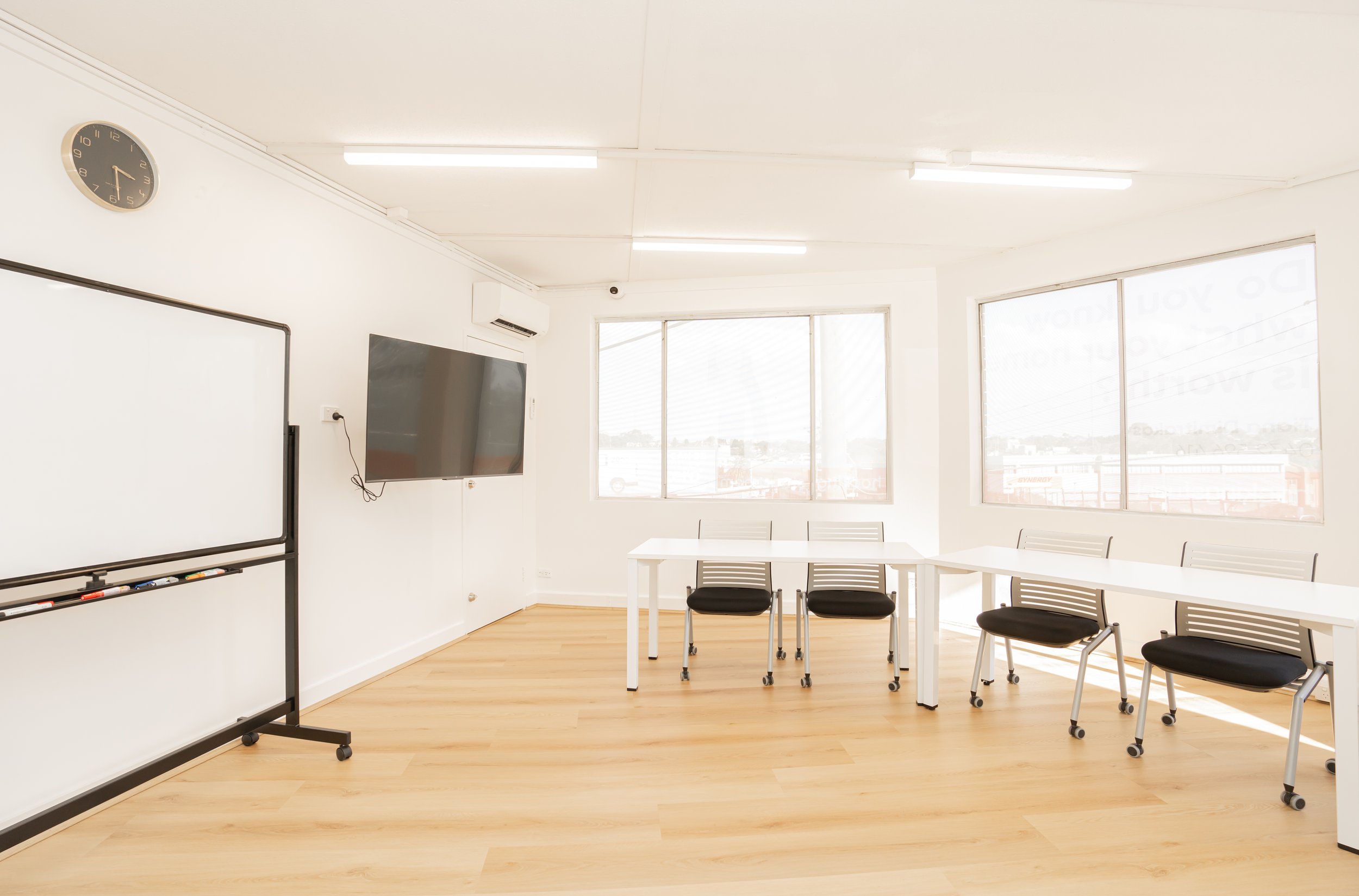 Empty classroom with white walls, large windows, a whiteboard on a black stand, a wall-mounted TV, a clock showing 1:00, white tables, black chairs, and wood flooring.