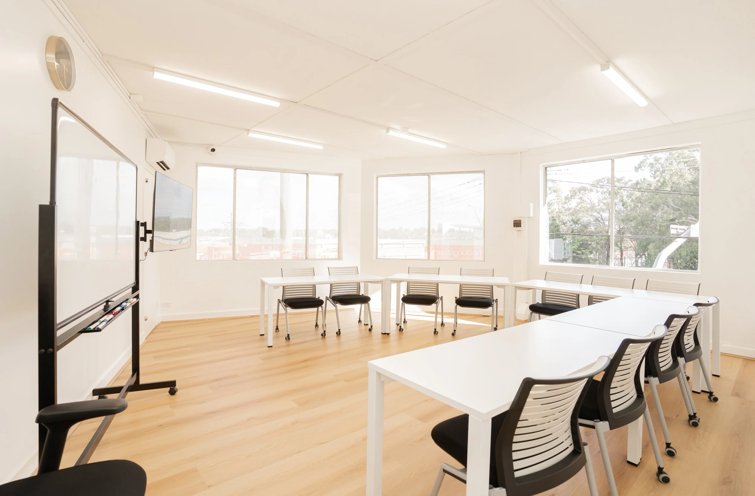 Empty classroom with white walls, large windows, white tables, black and white chairs, a whiteboard, a television, and a wall clock.