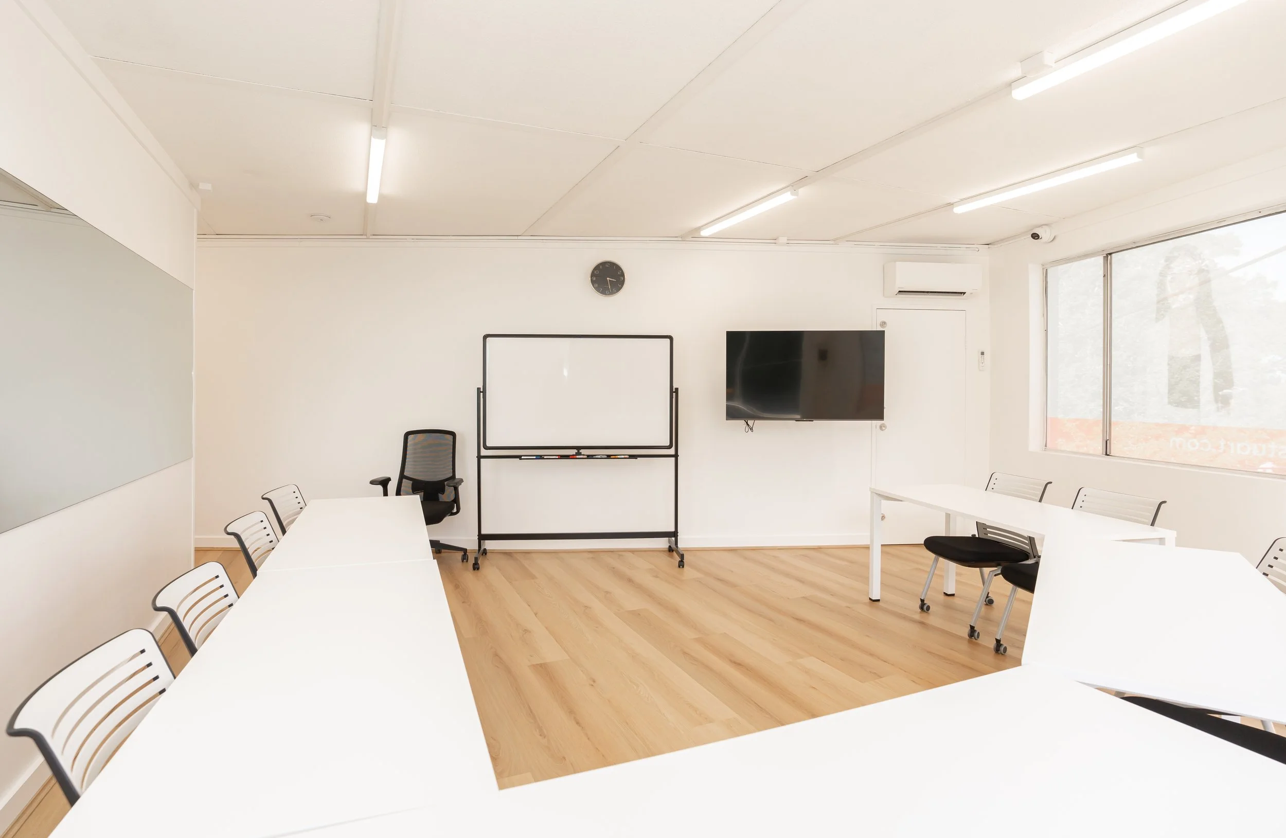 Empty modern conference room with white tables, chairs, whiteboard, wall clock, TV, and large window.