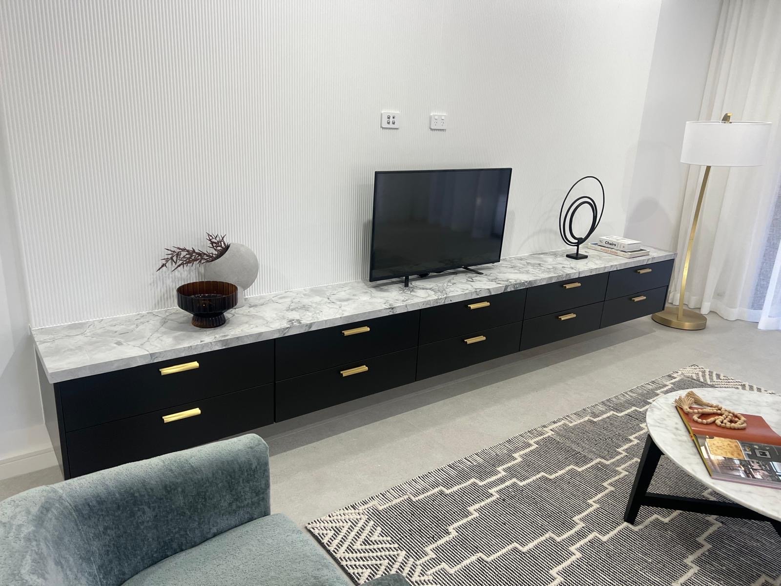 Modern living room with a black and white marble TV console, a flat-screen TV, decorative objects, a standing lamp, a patterned rug, and a coffee table with magazines.