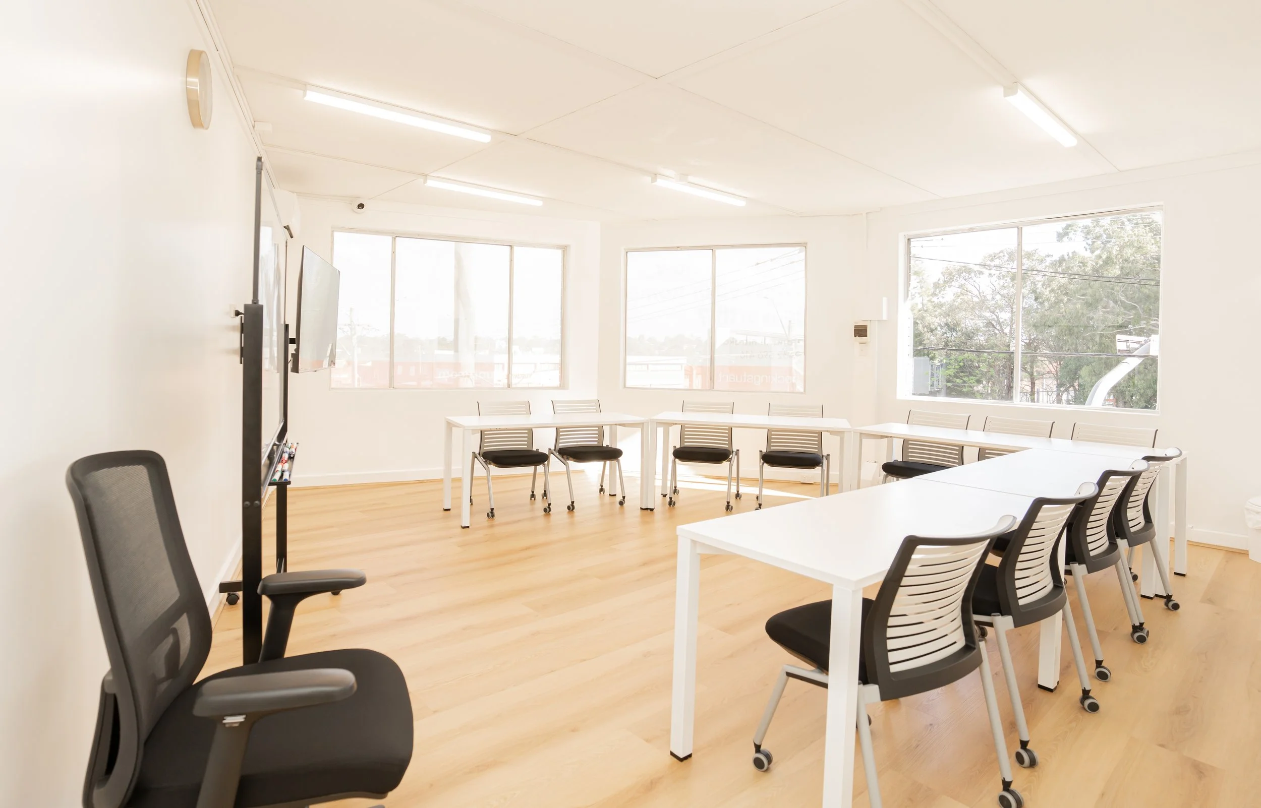 Empty classroom with white walls, wooden floor, white tables, black chairs, and large windows letting in natural light.