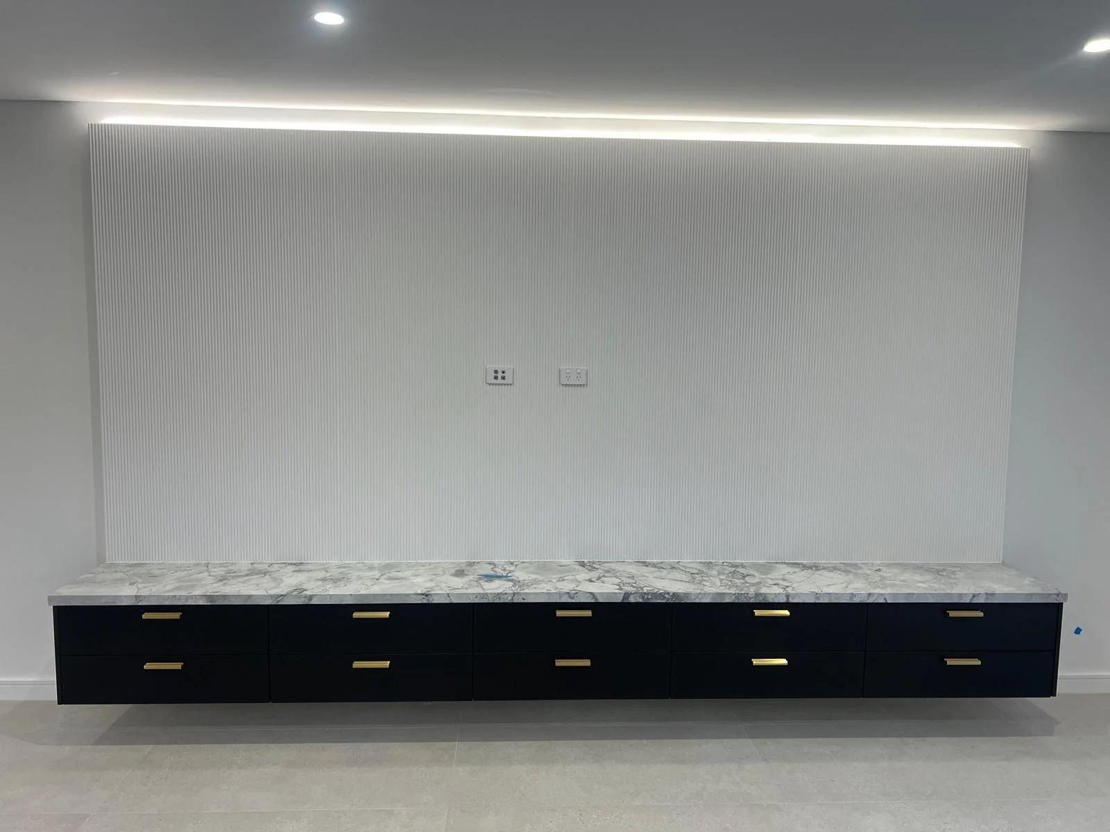 Minimalist interior with a white wall featuring vertical grooves, a marble countertop, and below it a black cabinet with gold handles.