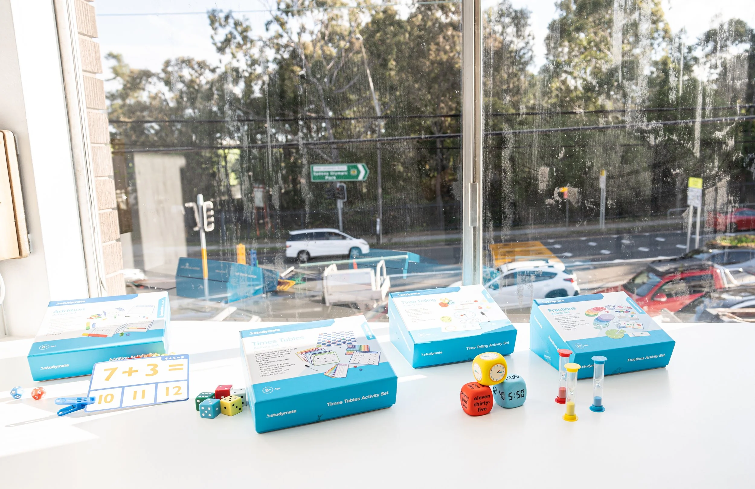 Educational activity sets on a white table by a window, including boxes labeled with math activities, dice, clocks, time cubes, and sand timers.