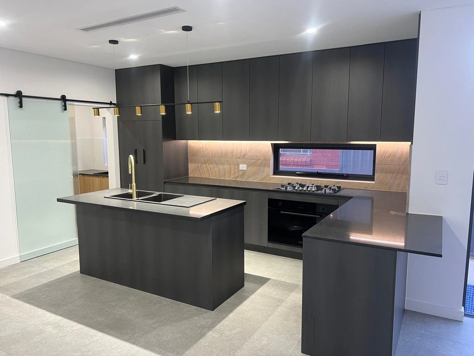 Modern kitchen with black cabinetry, a marble island with a sink and gold faucet, a built-in oven, a gas cooktop beneath a window, and pendant lighting.