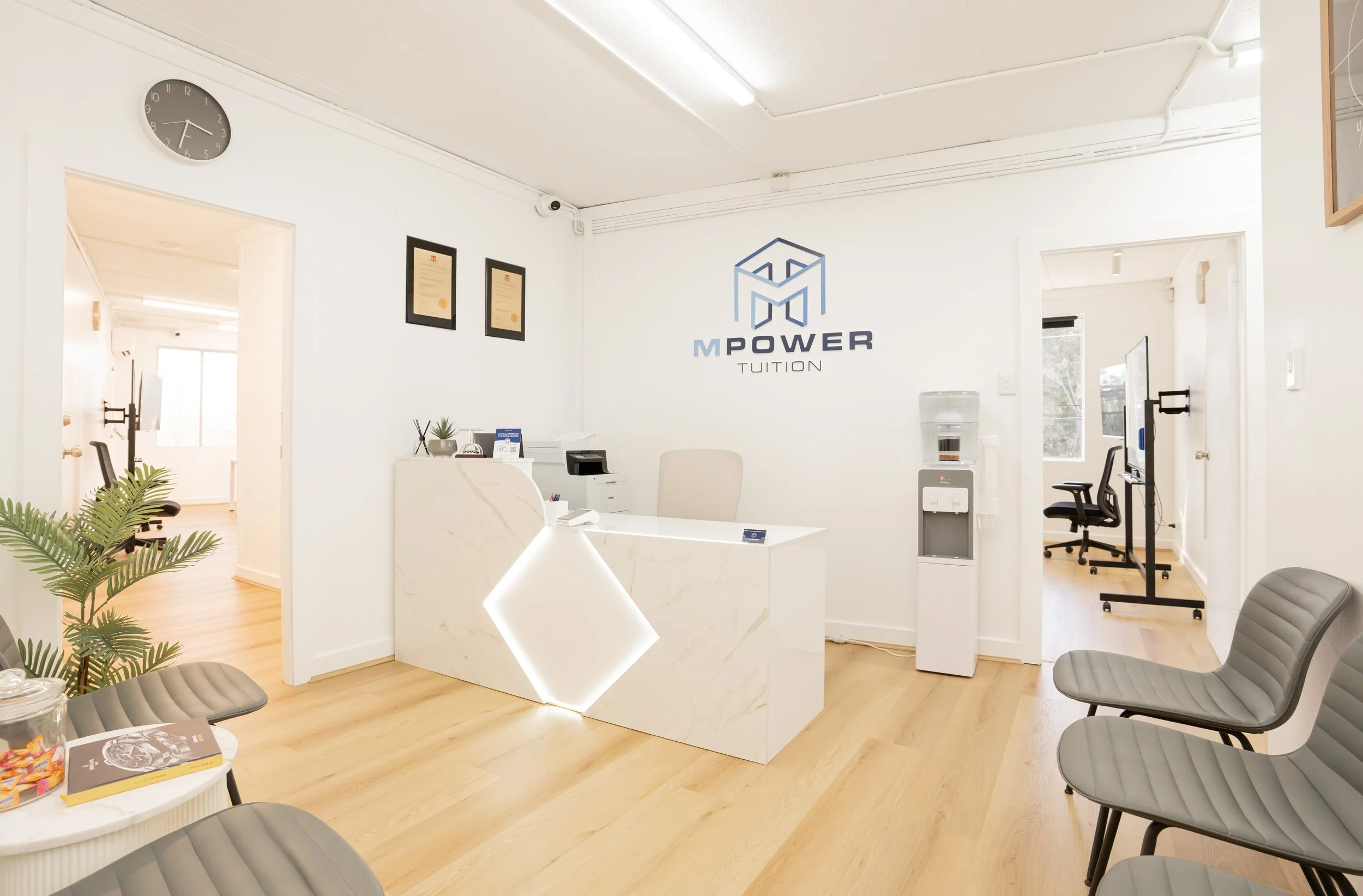 Modern reception area with white walls, wooden floors, a marble desk with under-lighting, chairs, water dispenser, water cooler, and framed certificates on the wall. The logo 'M Power Tuition' is on the wall behind the desk.