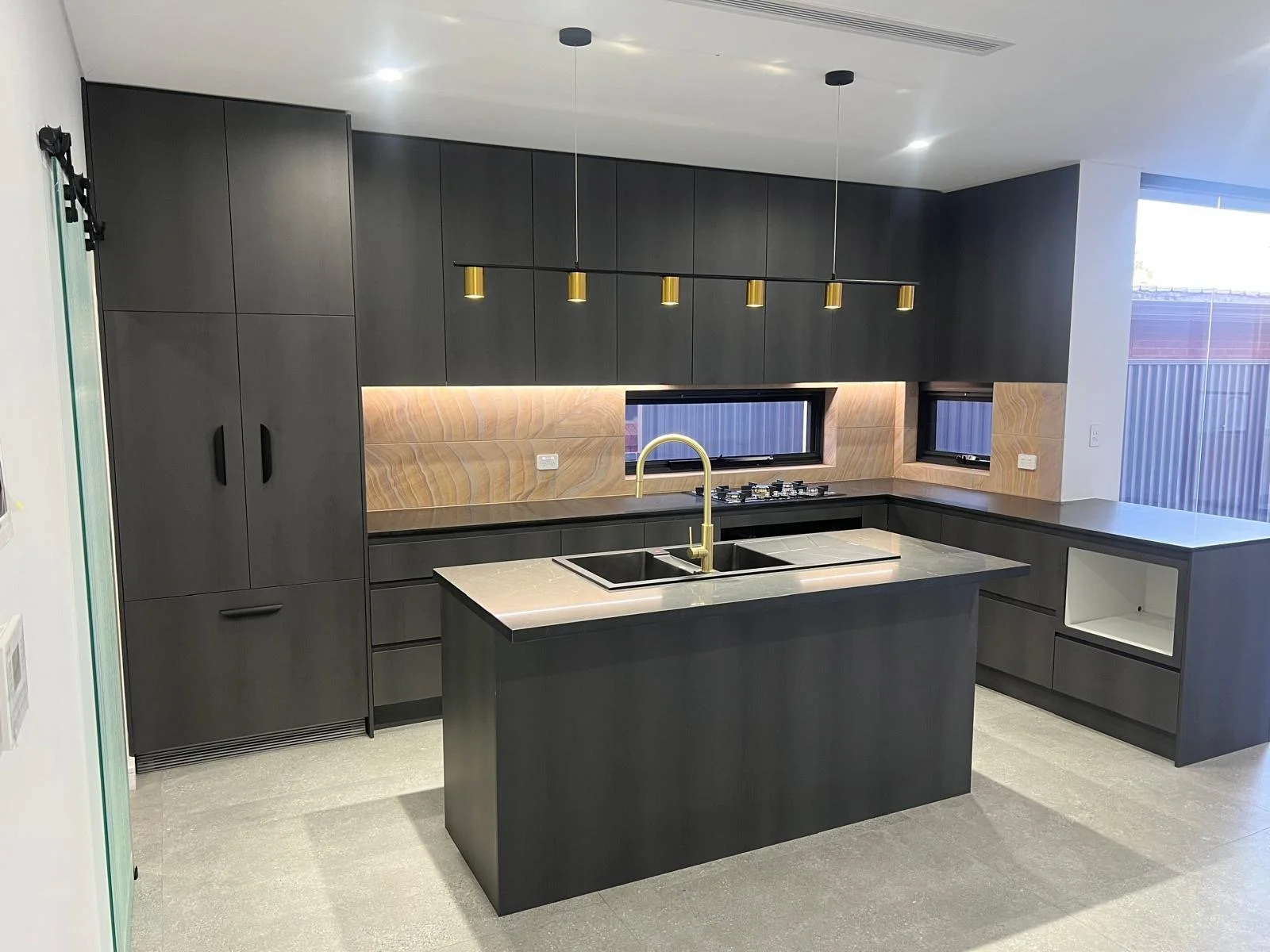 Modern kitchen with dark cabinets, a center island with a sink and golden faucet, two windows with black frames, under-cabinet lighting, and a gray countertop.