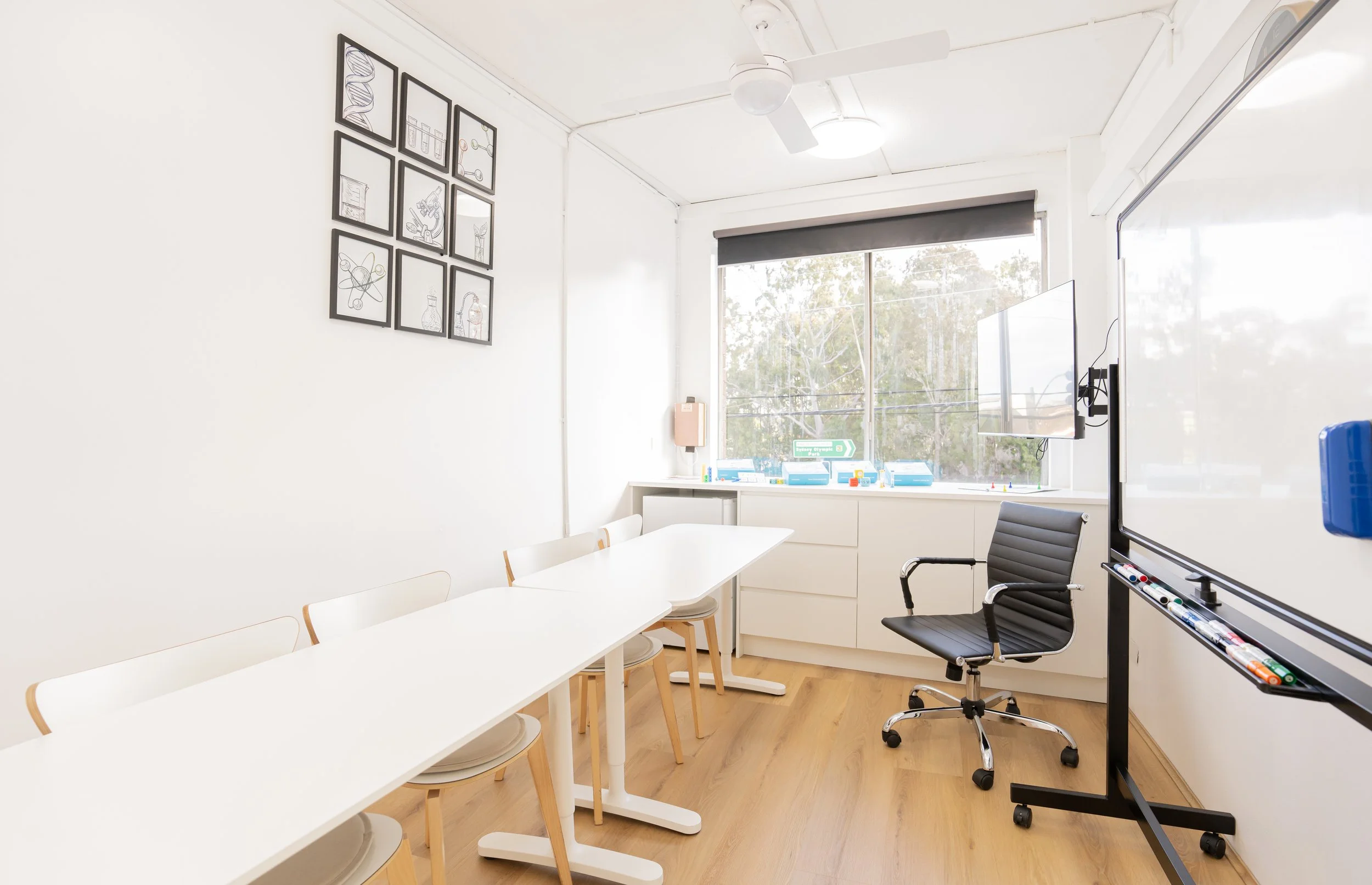 A bright, modern classroom with white walls, a white table, white and wooden chairs, a black office chair, a large window with a gray blind, a white cabinet, and a whiteboard with markers.