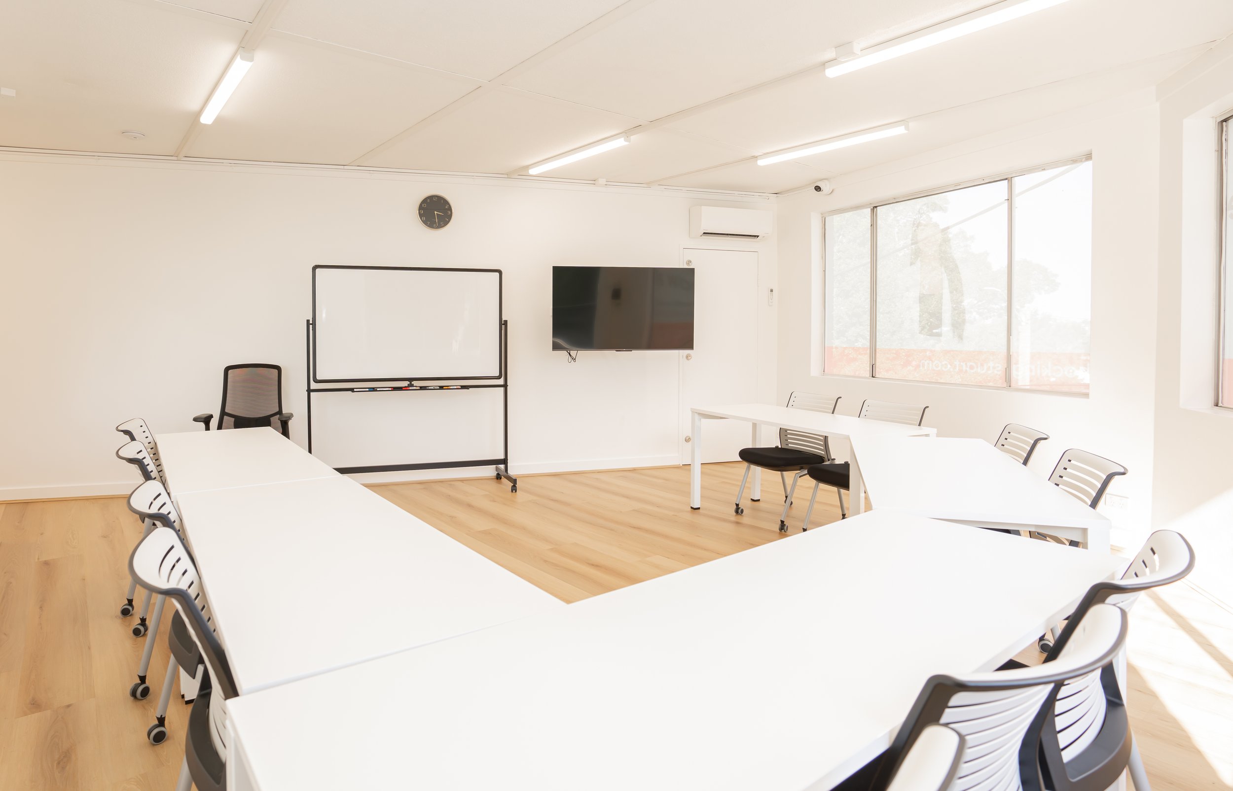 Empty classroom with white tables arranged in a U-shape, black chairs, a whiteboard, a wall-mounted television, a clock, and large windows letting in natural light.