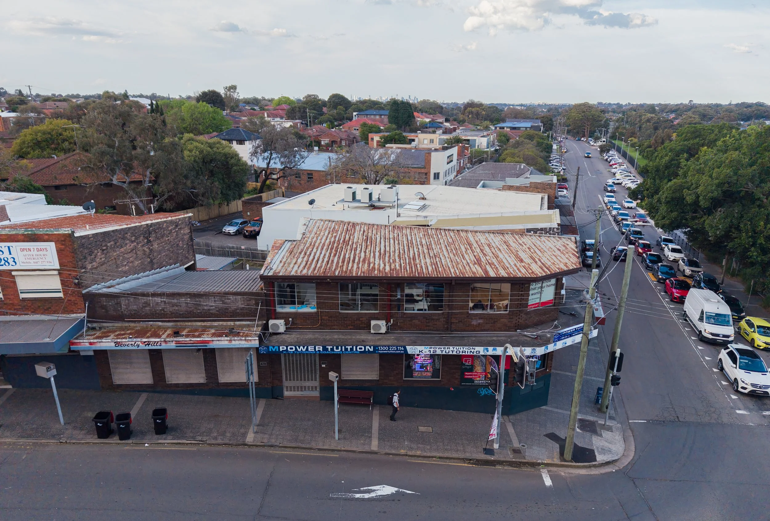 Aerial view of a small commercial building with a rusted metal roof, located at a street corner. The building has signs advertising tutoring and coaching services, and there are parking spaces along the street and behind the building. Several cars ar