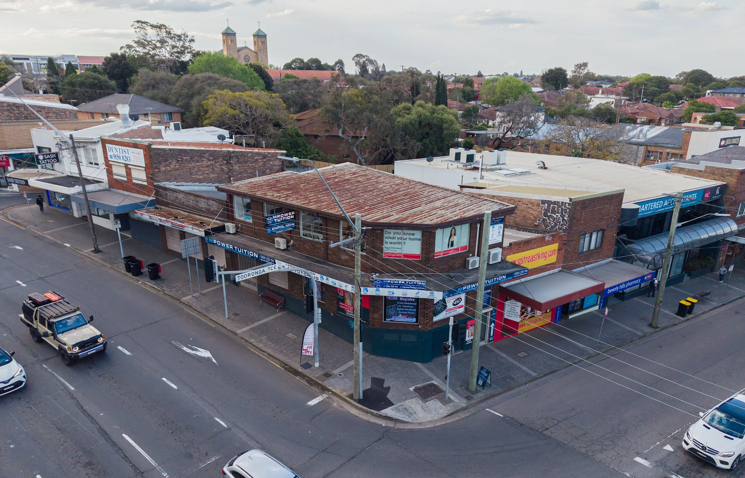 Aerial view of a corner building on a street with multiple signs and air conditioning units, surrounded by parked cars and trees, with houses and a church with twin towers in the background.