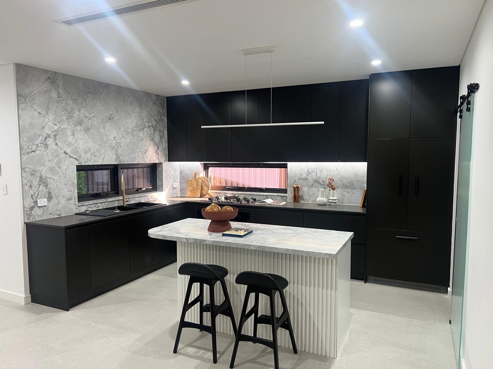 Modern kitchen with black cabinetry, marble countertops, and a marble backsplash. Includes a kitchen island with a marble top, two black barstools, and various decorative items.