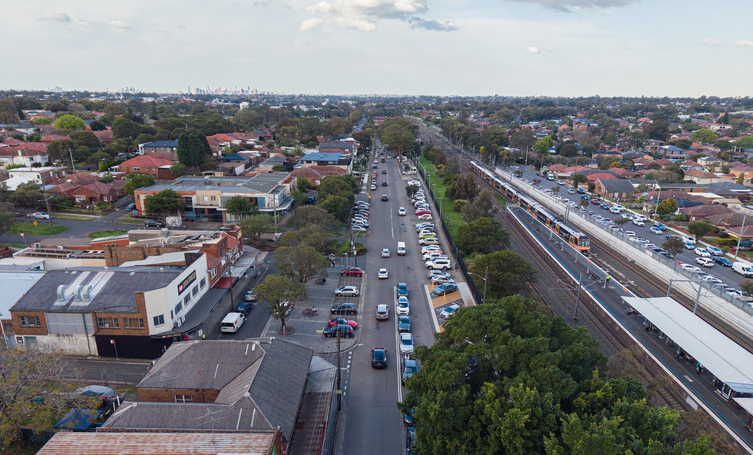 Aerial view of a residential and commercial area with a train passing on tracks and a central road with parked and moving cars, surrounded by trees and houses, city skyline visible in the distance.