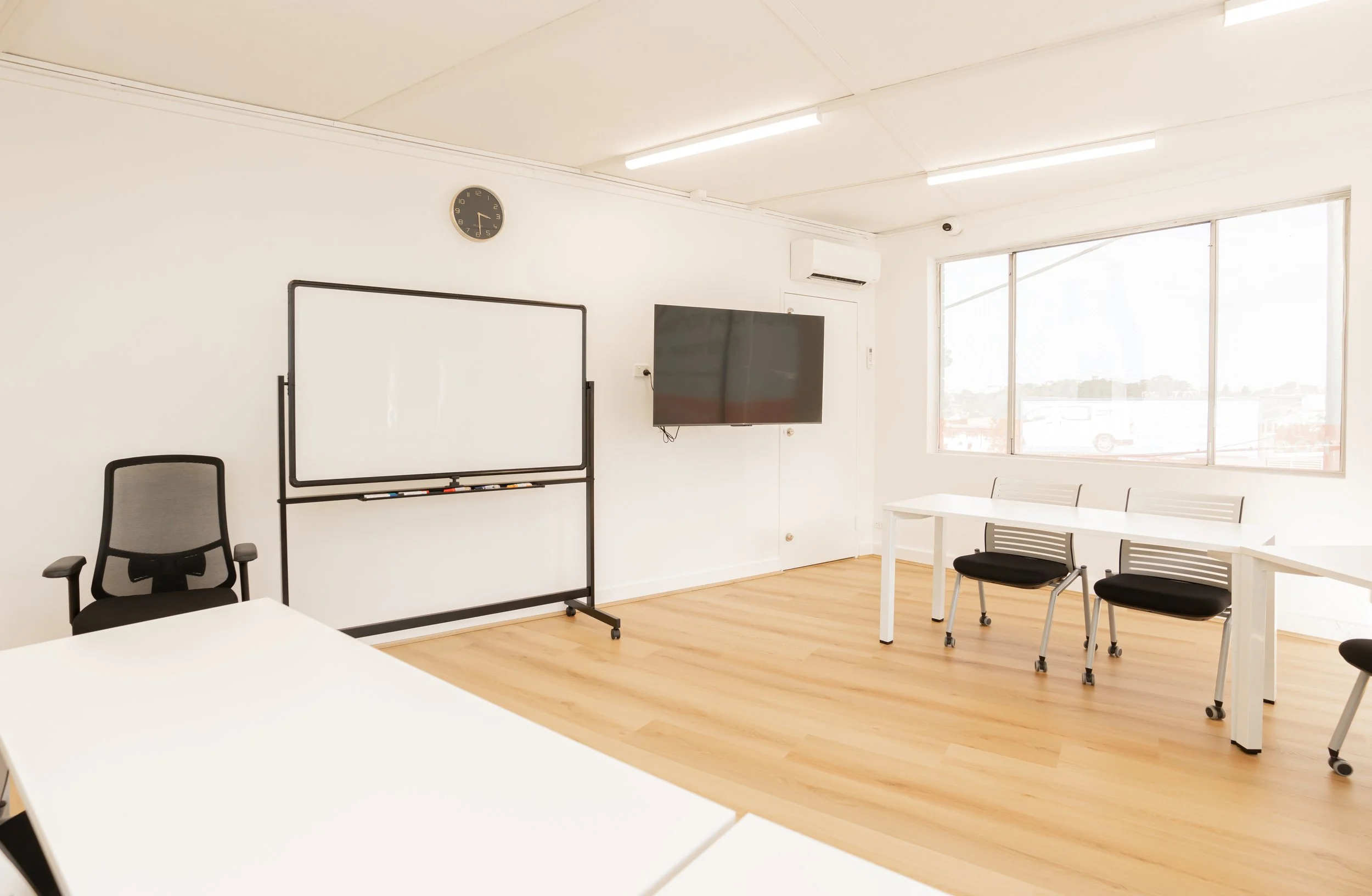 Empty classroom with white tables, black chairs, whiteboard, wall clock, flat-screen TV, and large window with a view outside.