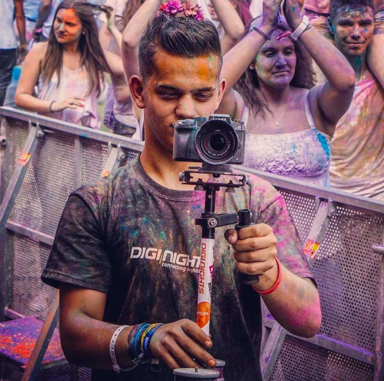 Junge bei einem Farbfestival mit einer Kamera auf einem Handstativ, umgeben von feiernden Menschen.
