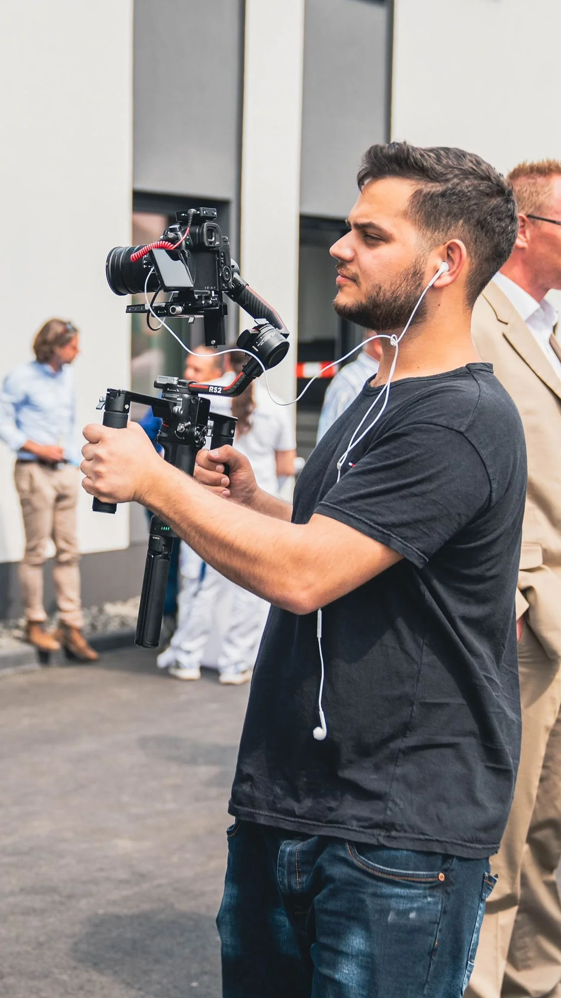 Junger Mann filmt mit einer Kamera auf einem Gimbal, trägt Ohrhörer, im Hintergrund sind andere Erwachsene in einem urbanen Umfeld.