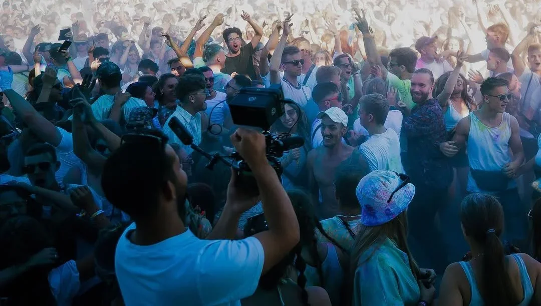 Eine große Menschenmenge bei einem Musikfestival oder Club, viele tanzen und haben Spaß, einige tragen Sonnenbrillen, während andere ein Kamerateam filmen.