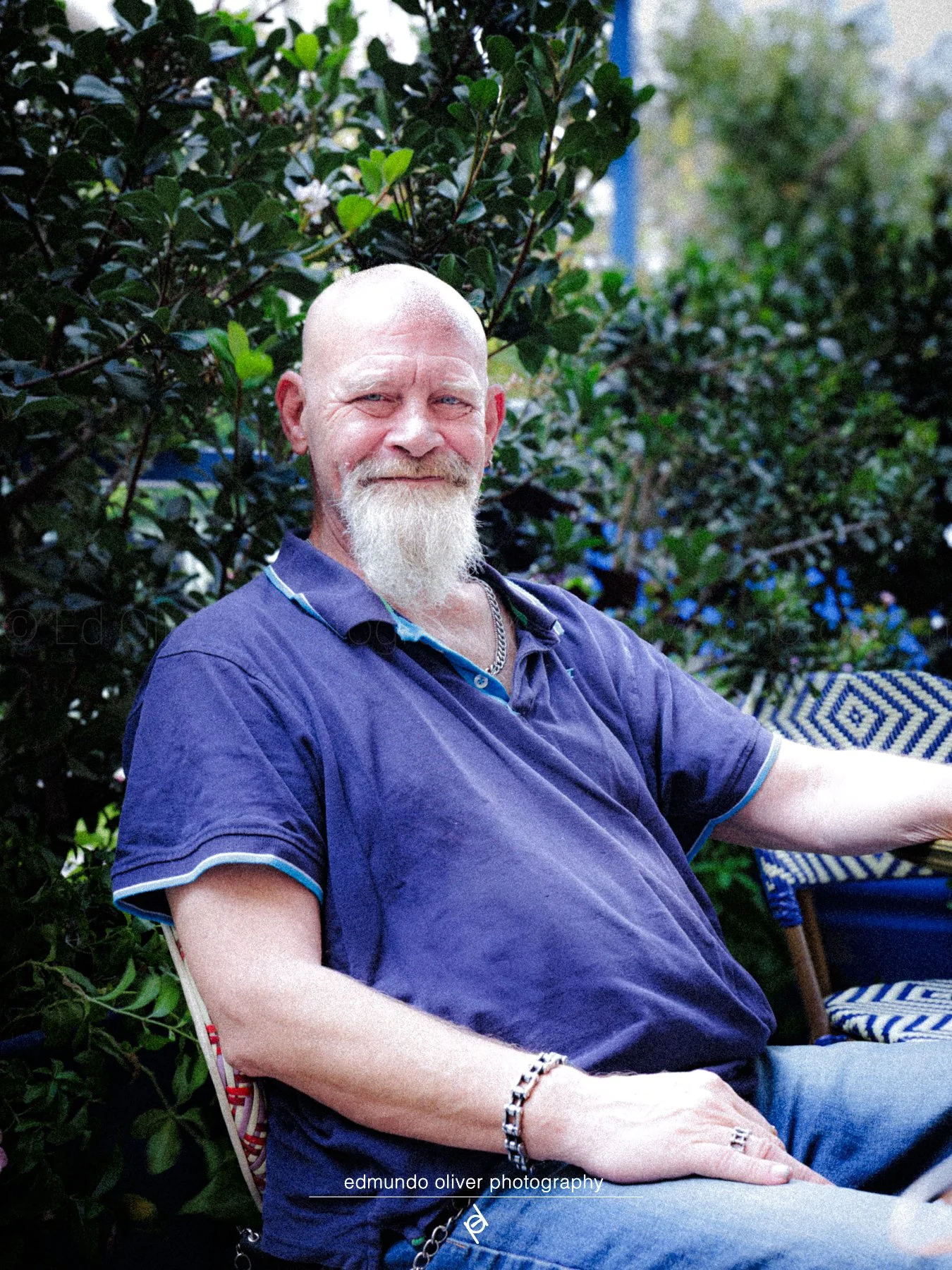 A man with a white beard and bald head sitting outdoors in front of green bushes, wearing a purple polo shirt with a chain necklace and bracelets, smiling slightly.