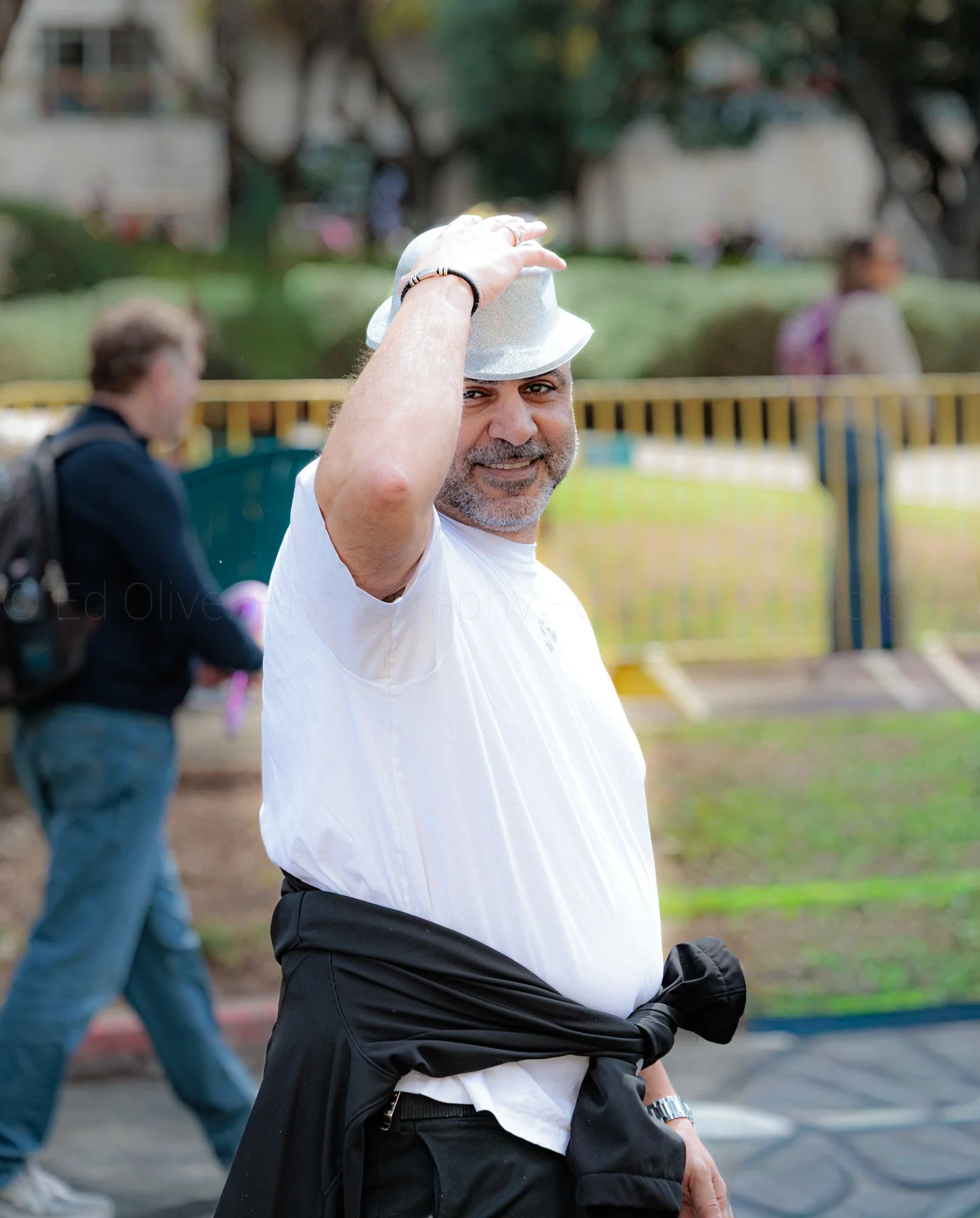 A smiling man wearing a white shirt, black pants, and a silver hat, standing outdoors with a blurred background of other people and trees.