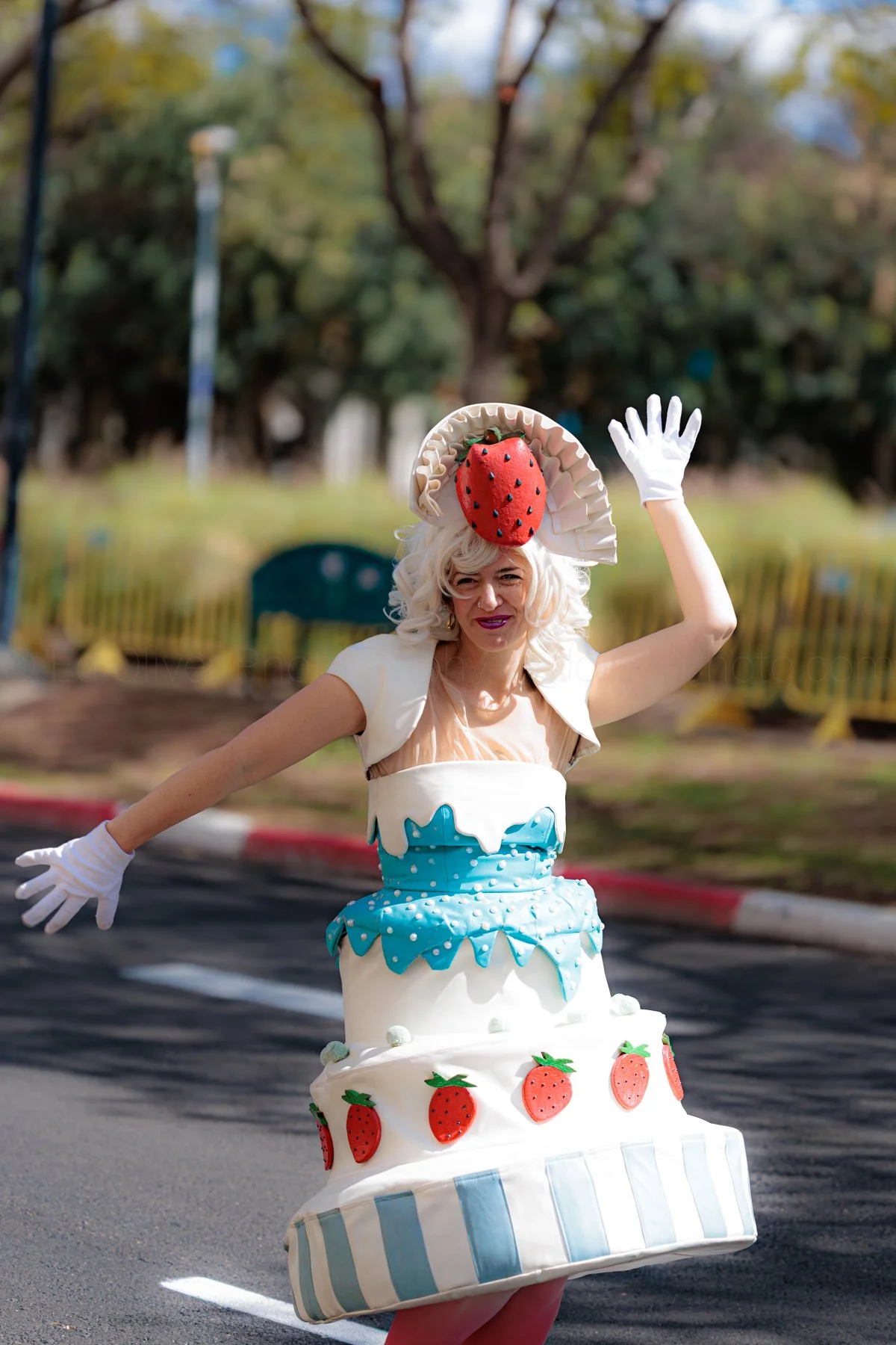 Person dressed in cake costume featuring strawberries, waving outdoors with trees and park in background.