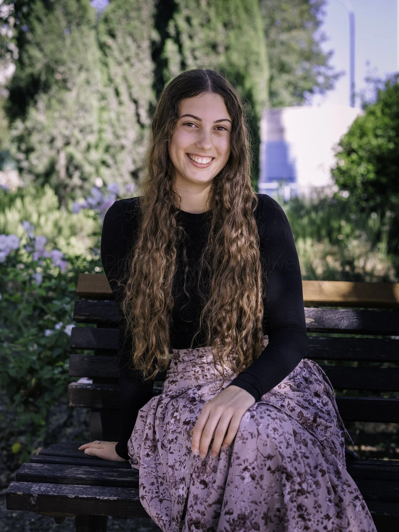 A young woman with long, wavy hair sitting on a park bench outdoors.