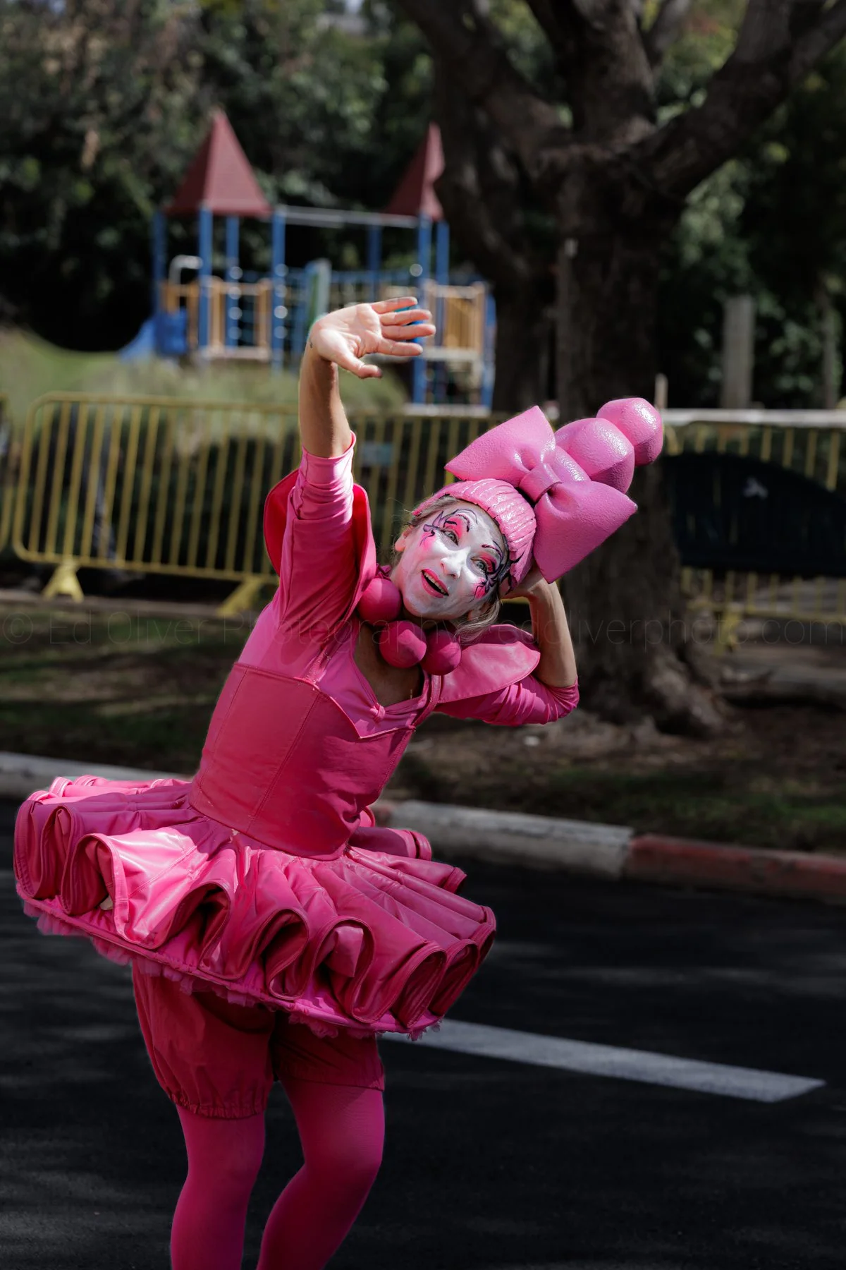 A person dressed in a vibrant pink costume with elaborate makeup and a headpiece, performing on a street with goal posts and playground equipment in the background.