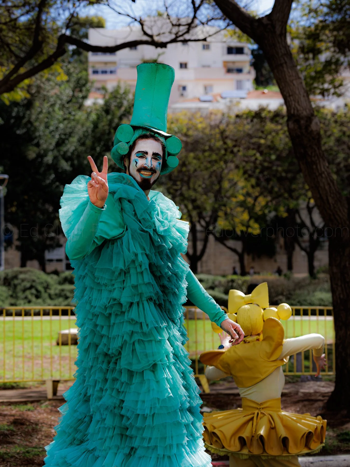 Performer dressed as a clown or theatrical character in a teal ruffled costume with a tall teal hat, painted face, and holding up a peace sign in an outdoor park with trees and a building in the background.