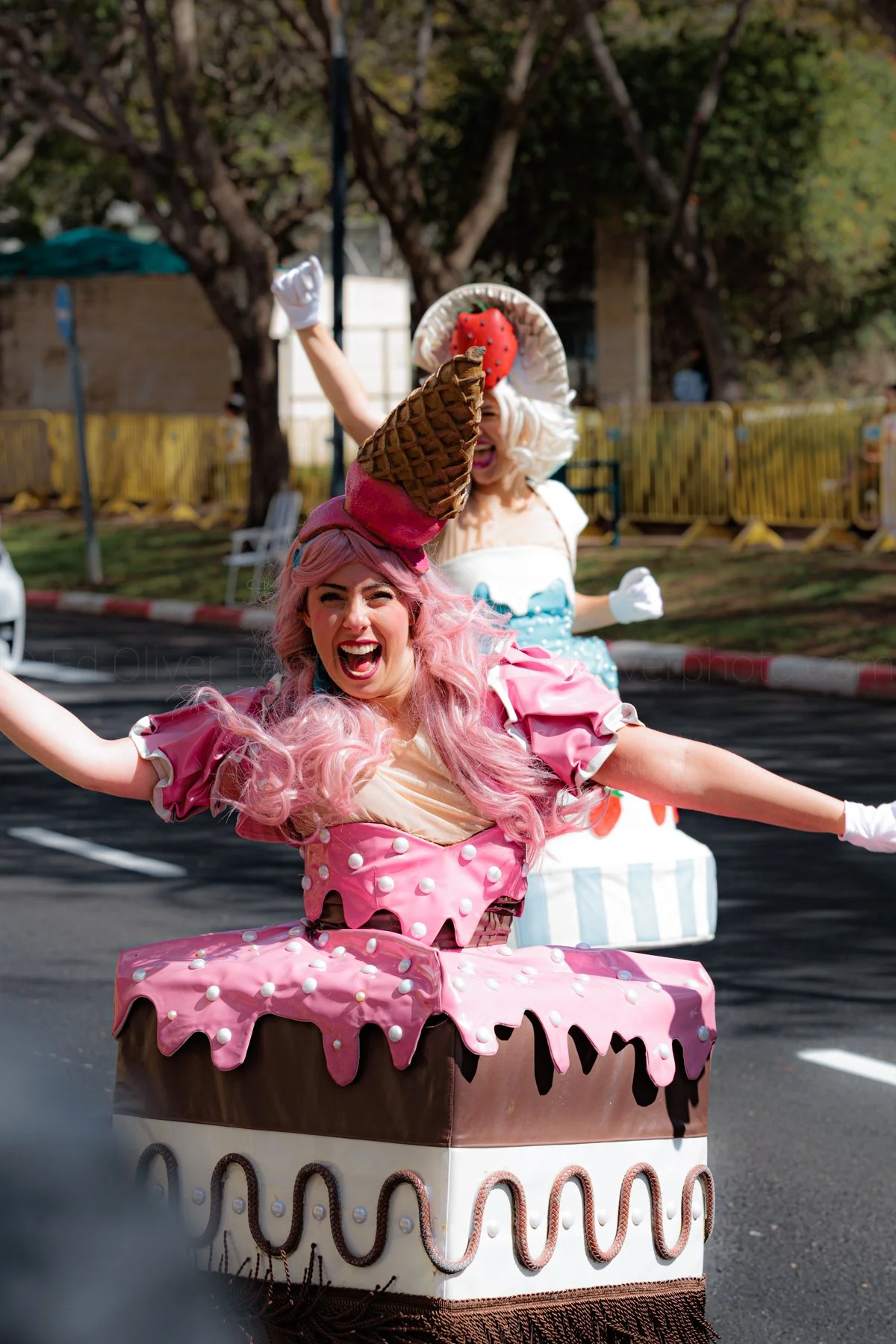 Two women dressed as cake and ice cream characters celebrating outdoors, with trees and a yellow fence in the background.