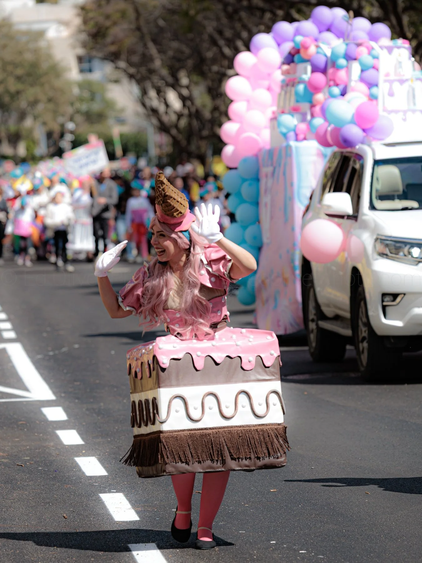 A woman dressed in a cake-themed costume walking in a parade, with a pink crown and ice cream cone on her head, and a large cake-shaped skirt; she is smiling and waving among a crowd and decorated float with balloons.