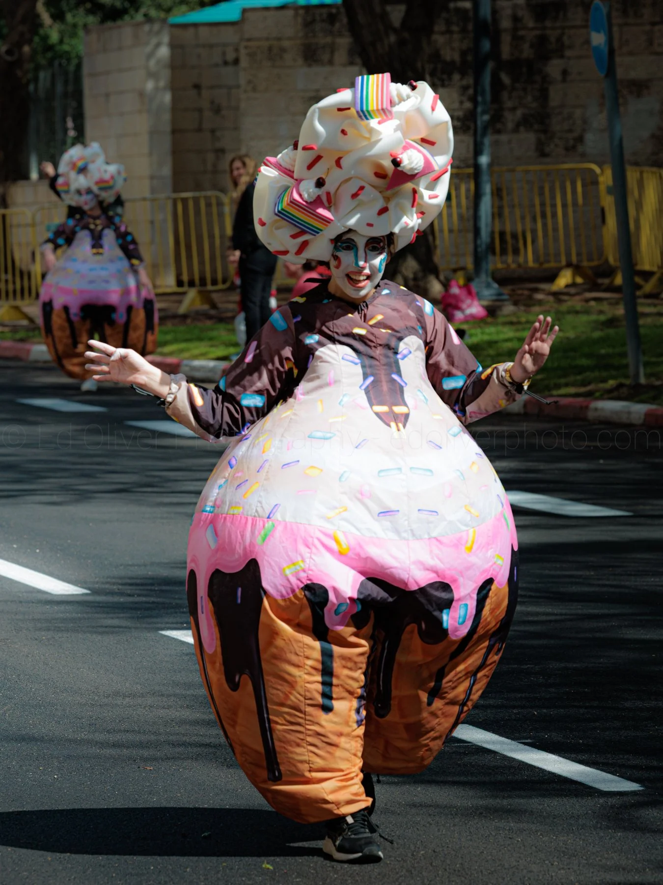 Person dressed in a cupcake costume with colorful sprinkles, a large whipped cream and cherry headpiece, and face makeup shaped like a cupcake, walking on the street during a parade.
