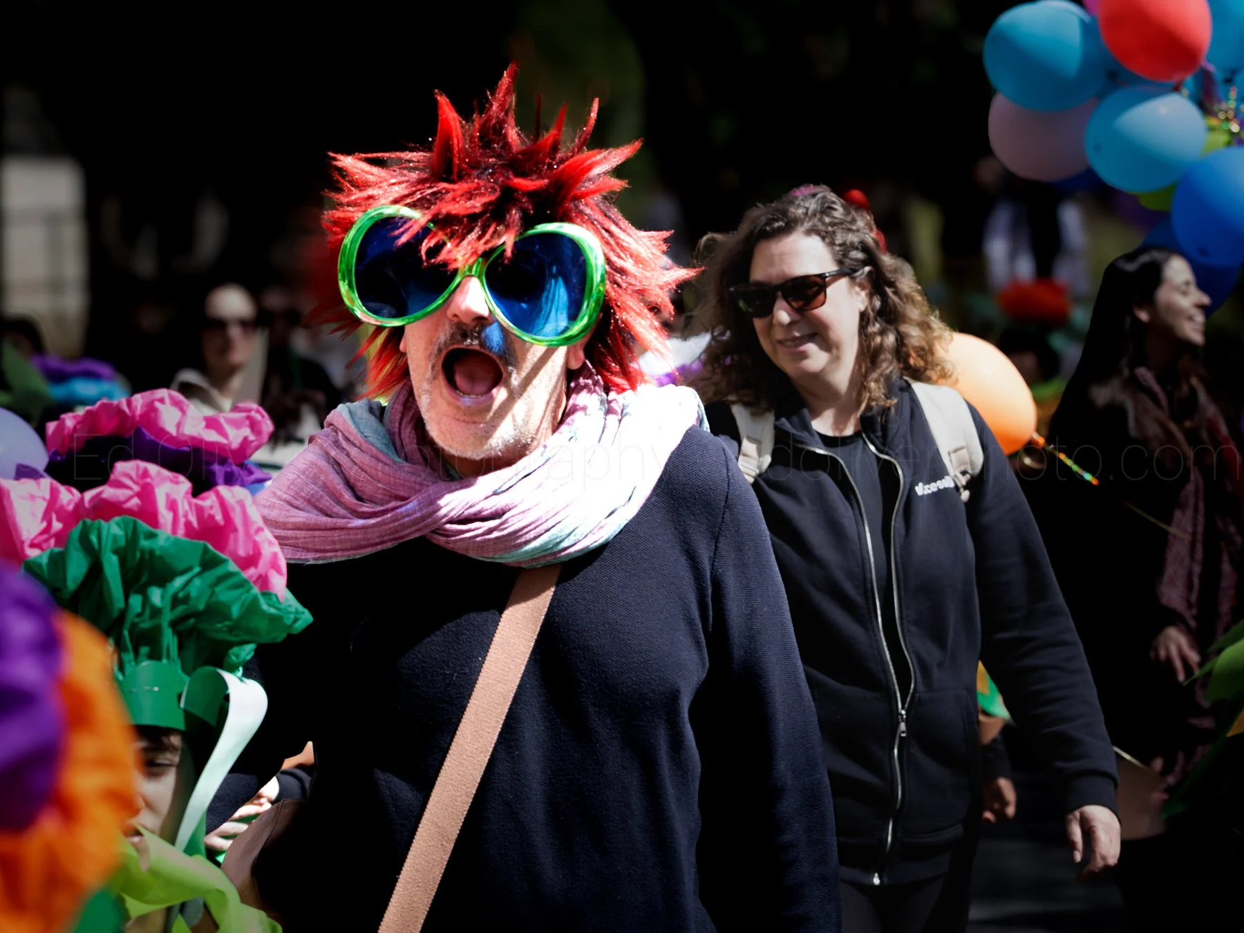 A person with red spiked hair and large colorful sunglasses is protesting and shouting. There is a woman in sunglasses and a black jacket smiling nearby. Bright balloons and flowers are visible in the background.