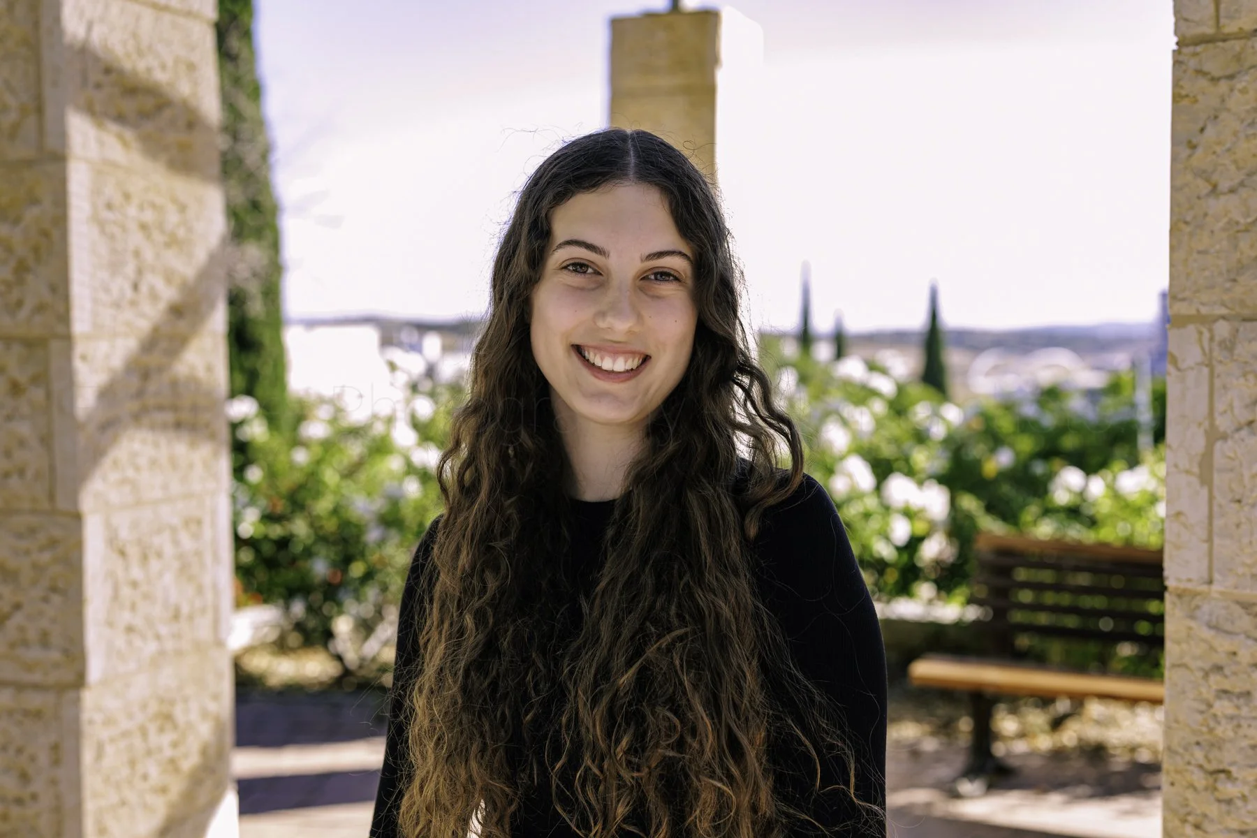 A young woman with long, curly brown hair and a black shirt, smiling outdoors between stone columns with greenery and a bench in the background.