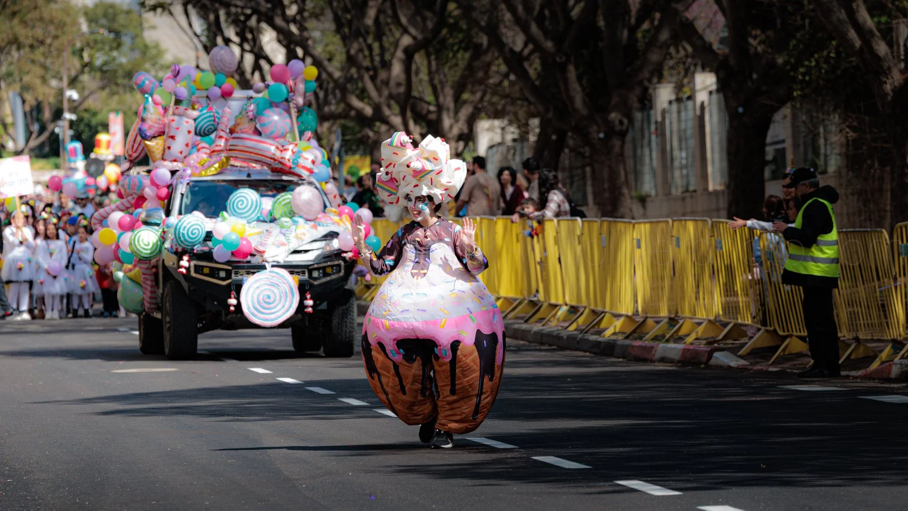 A person dressed in a colorful cake costume with pink frosting and ice cream details, walking in a parade. In the background, a decorated vehicle covered with balloons and candies, and spectators behind yellow barriers.