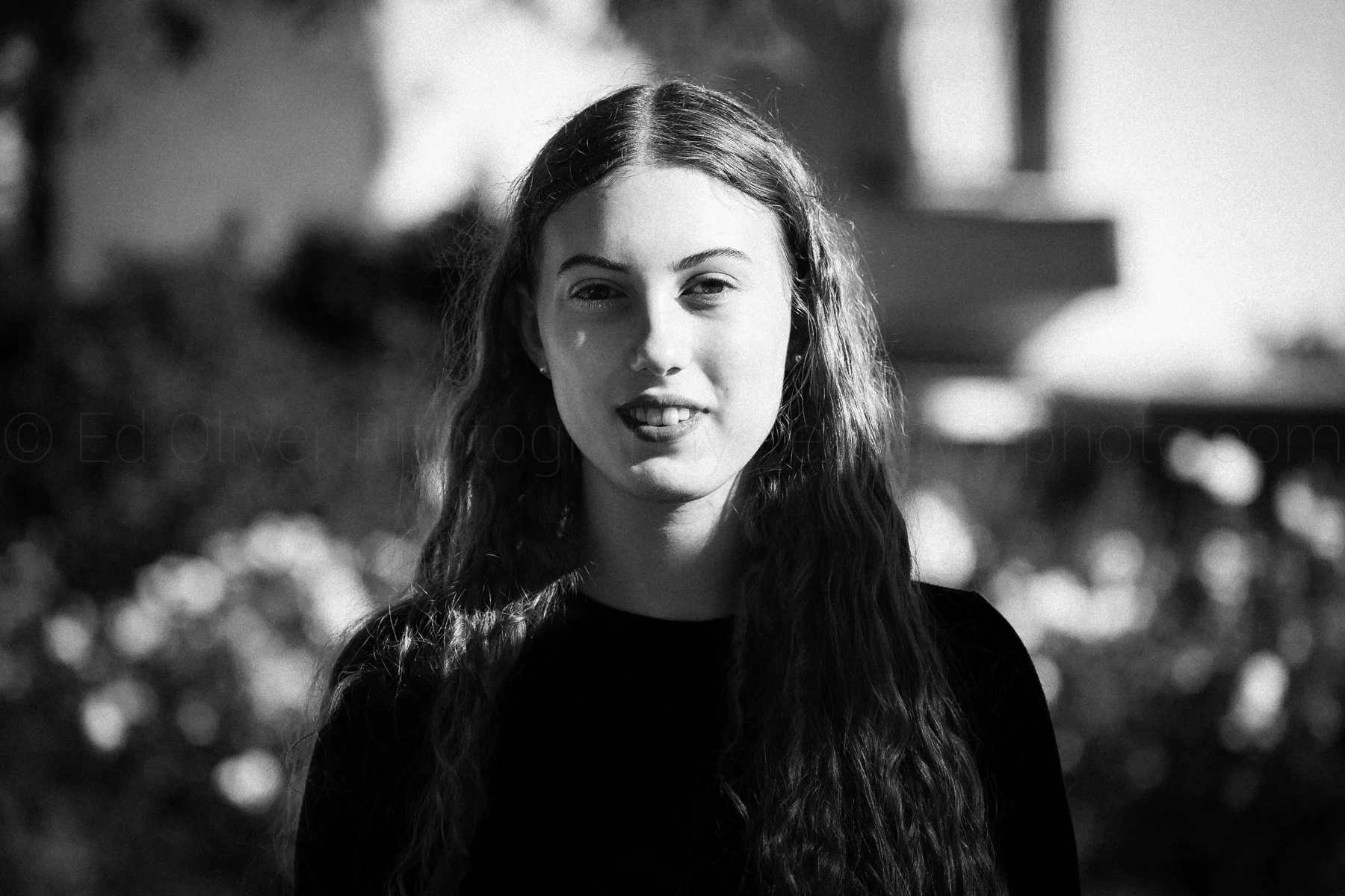 Black and white portrait of a young woman with long, wavy hair, smiling slightly, standing outdoors with blurred background foliage.