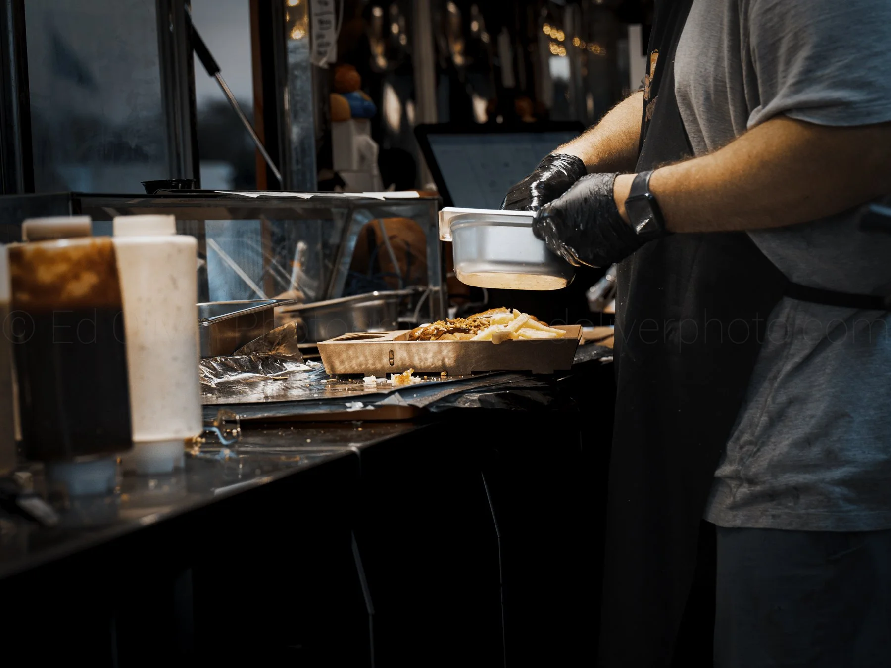 Person wearing a gray T-shirt and black gloves preparing food at a food truck or outdoor food stand, with various condiments and food items visible on the countertop.