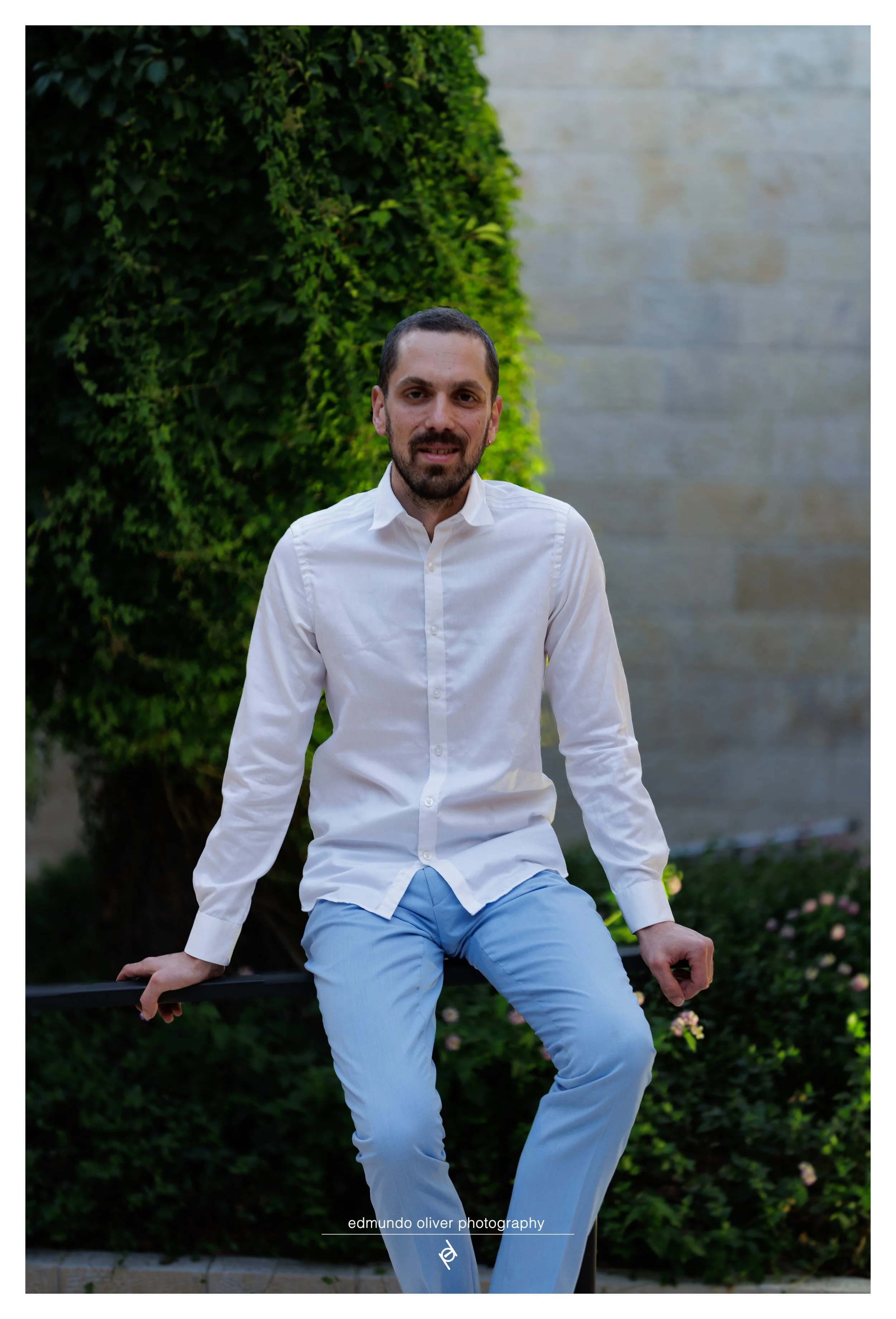 Portrait of a man with short dark hair and a beard, dressed in a white button-up shirt and light blue pants, sits on a park bench in front of greenery and a concrete wall.